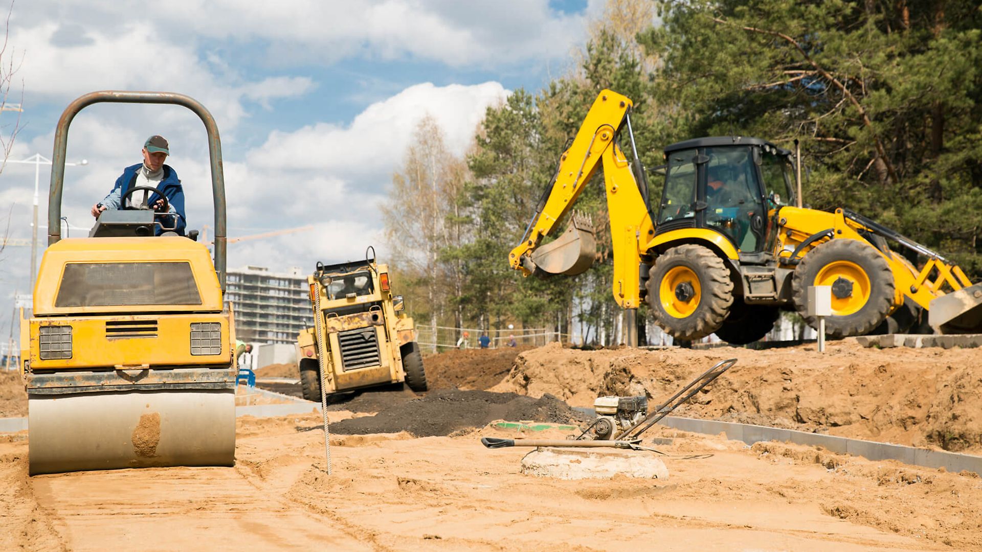 Auf dem Bild stehen 3 Maschinen. Ein Bagger, eine Straßenwalze und ein weiteres Fahrzeug. Ein Mitarbeiter fährt mit der Walze 