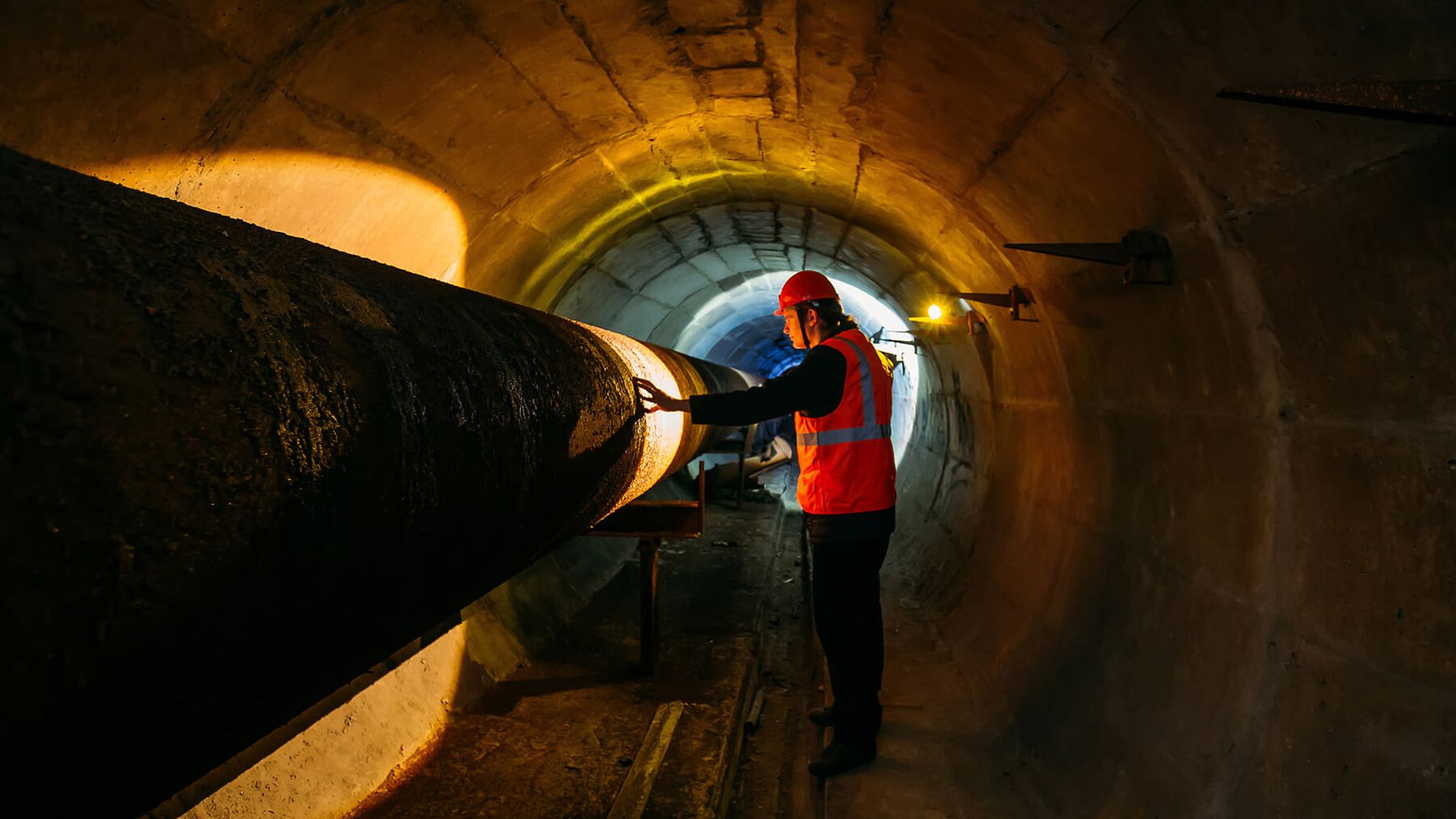 Eine Person mit Schutzweste und Helm steht in einem dunklen Tunnel. Er beleuchtet mit einer Arbeitslampe einen Teil eines Rohres