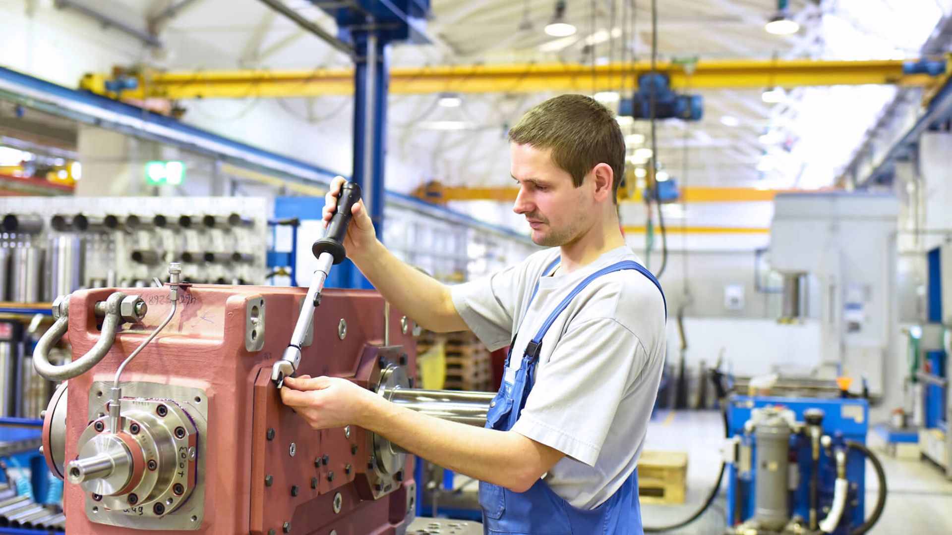 Ein Mann montiert eine Maschine. Im Hintergrund ist eine Halle mit mehreren Maschinen zu erkennen.