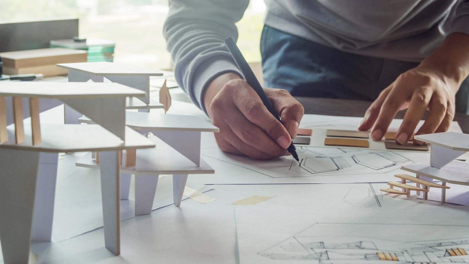 Es sind Architekturmodelle und Blätter mit technischen Zeichnungen zu sehen. Eine Person zeichnet gerade auf einem Blatt.