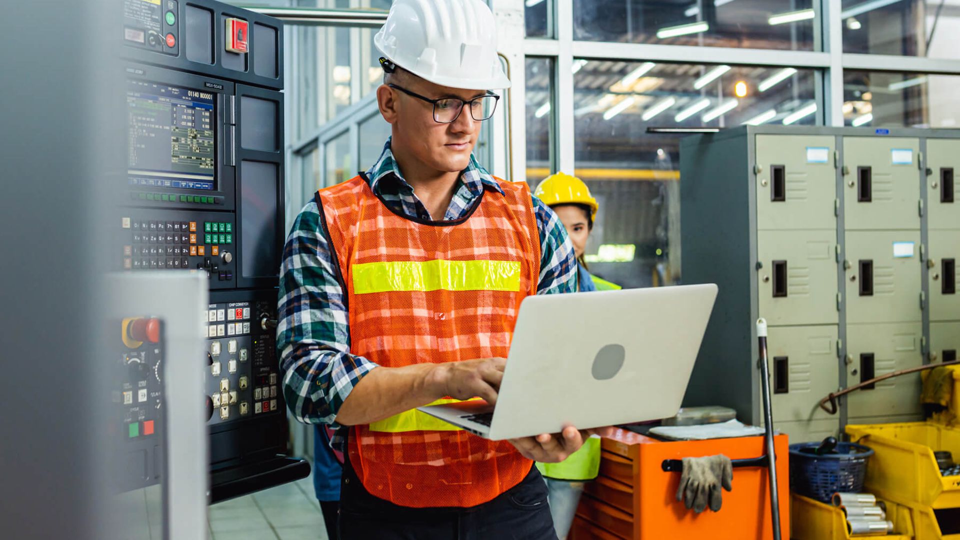 Ein Mann mit einem Laptop in der Hand steht vor einer großen Schalttafel mit vielen Knöpfen und Bildschirmen.