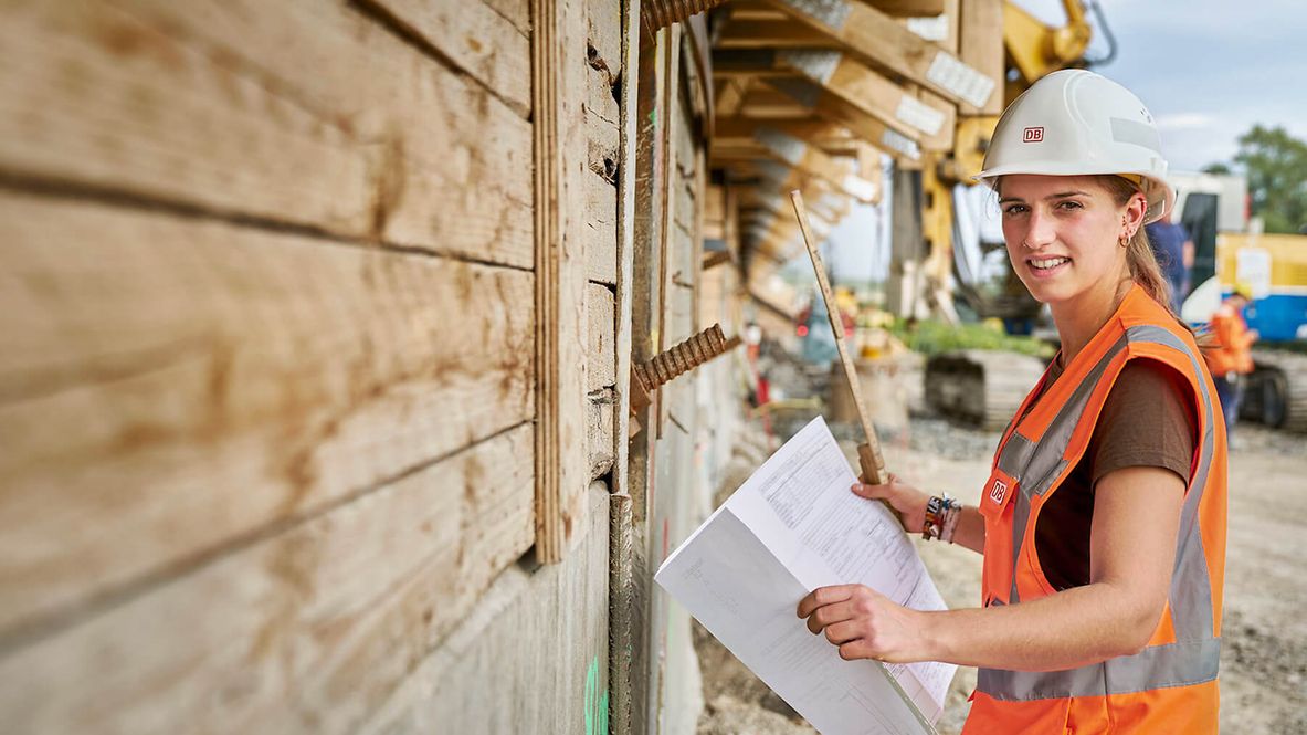 Eine Frau steht vor einem Gebäude aus Holz. Sie hat ein Plan in der Hand. Außerdem trägt sie Schutzkleidung.