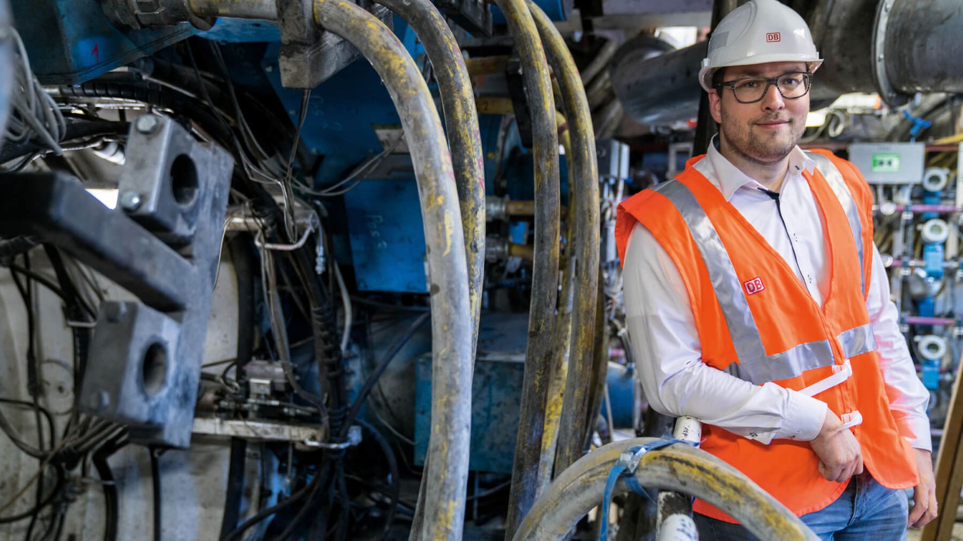 Ein Mann mit Schutzweste und Helm steht neben vielen Rohren und lächelt in die Kamera.
