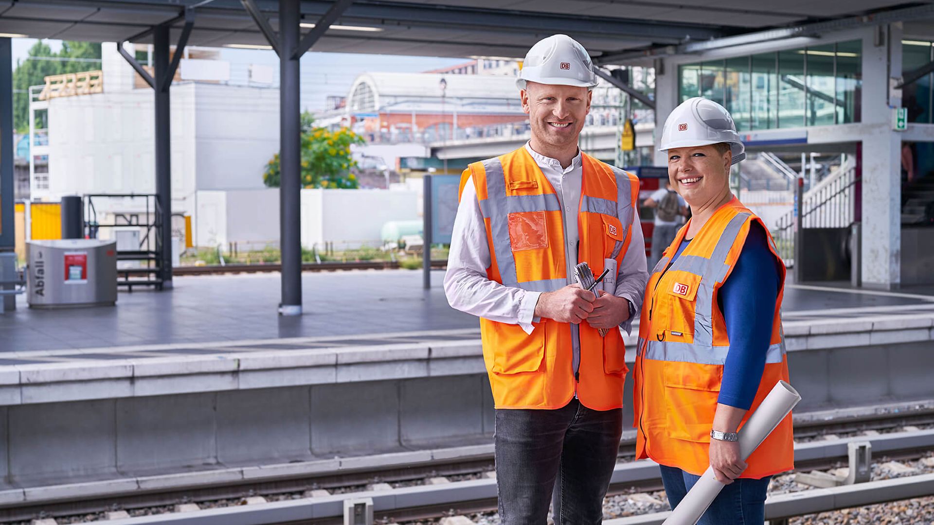 Zwei Personen mit Schutzwesten und Helmen stehen an einem Bahngleis. Im Hintergrund sieht man die Gleise