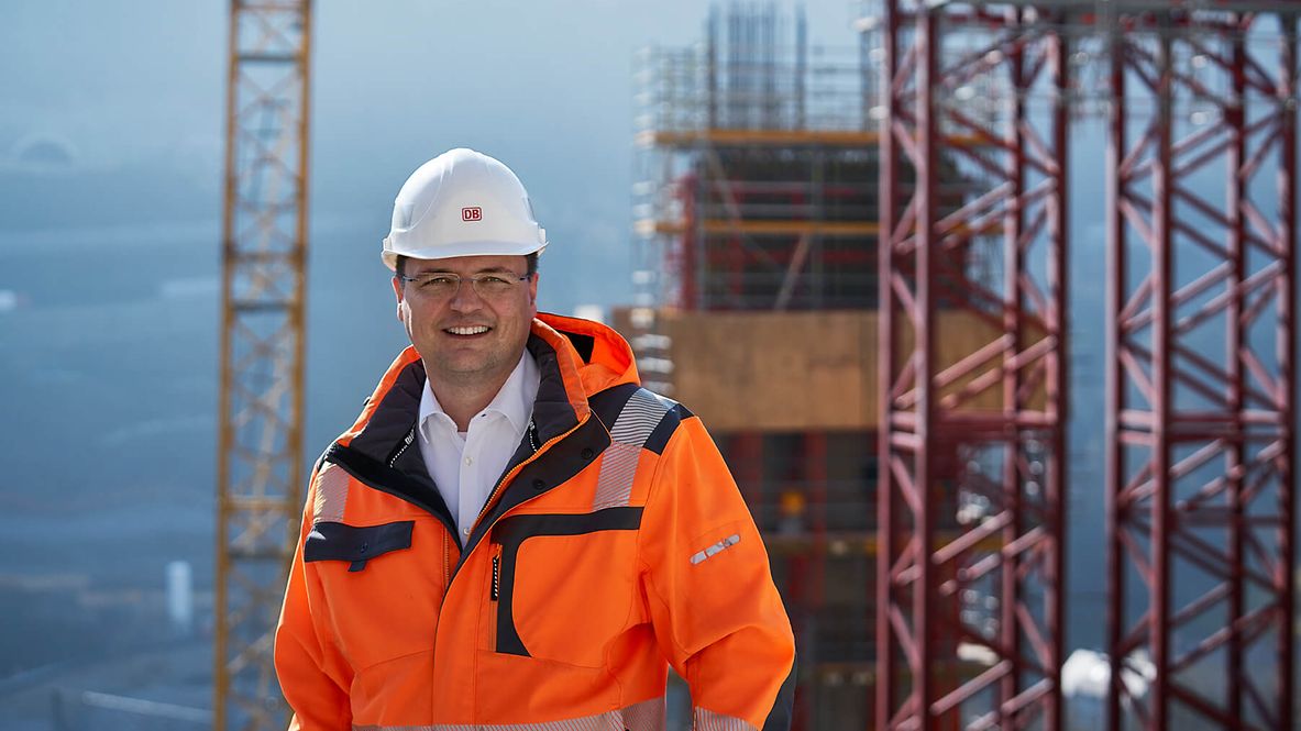 Ein Mann mit Neonfarbener Jacke und Schutzhelm lächelt in die Kamera. Im Hintergrund ist eine Baustelle zu sehen mit Kränen.