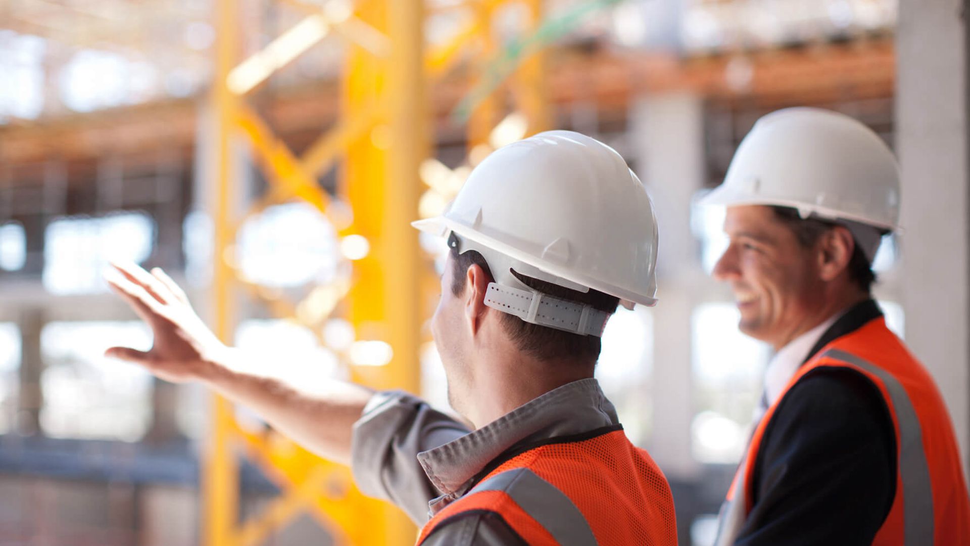 Zwei Bauleiter stehen vor einer Baustelle. Die eine Person streckt den Arm aus. Beide tragen Schutzhelm und Schutzwesten.