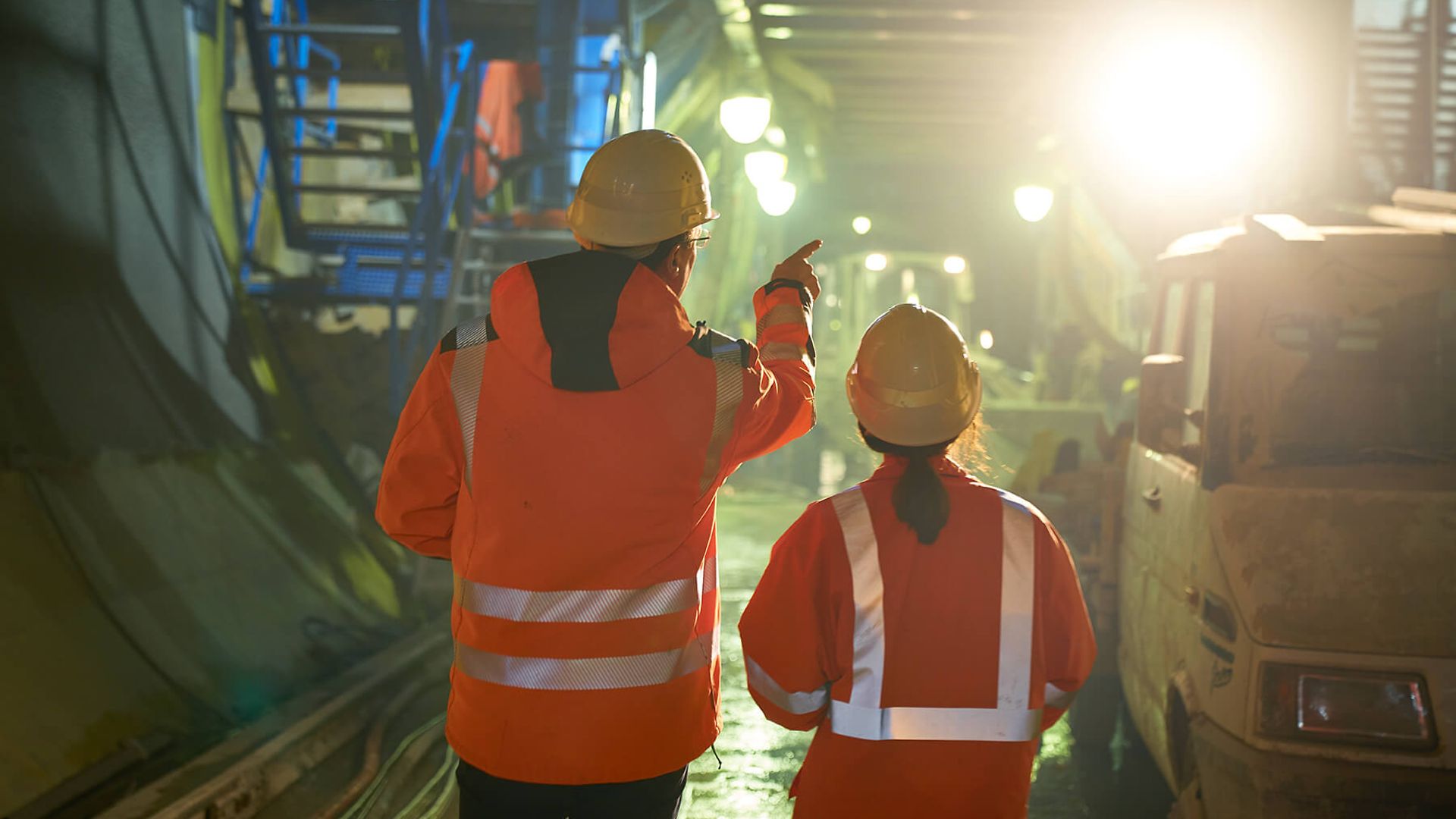 2 Personen mit Warnweste und Helm stehen auf einer Baustelle die im Untergrund ist. Die eine Person erklärt der anderen etwas.