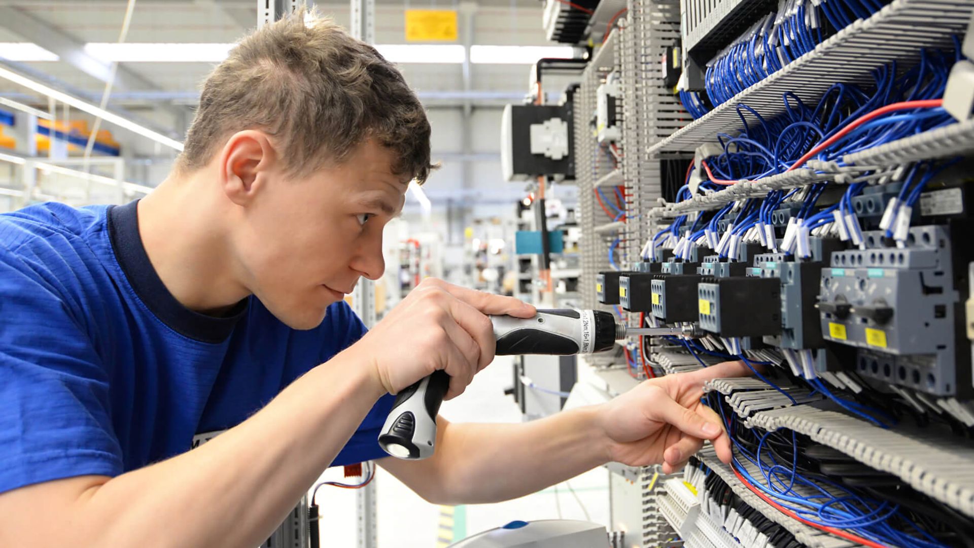Ein junger Mann schraubt mit einem Akkuschrauber Sicherungen in einem Schaltkasten fest.