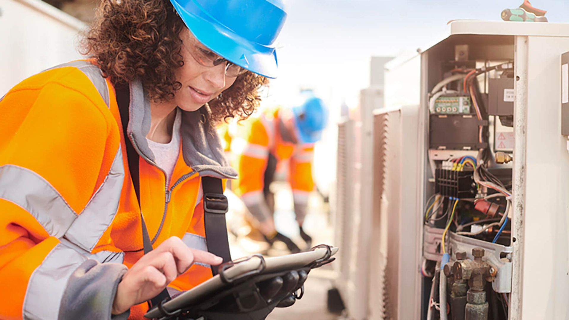 Eine Frau in Arbeitskleidung in Schutzhelm hockt vor einem Kasten in dem sich Kabel befinden und tippt auf einem Tablet.