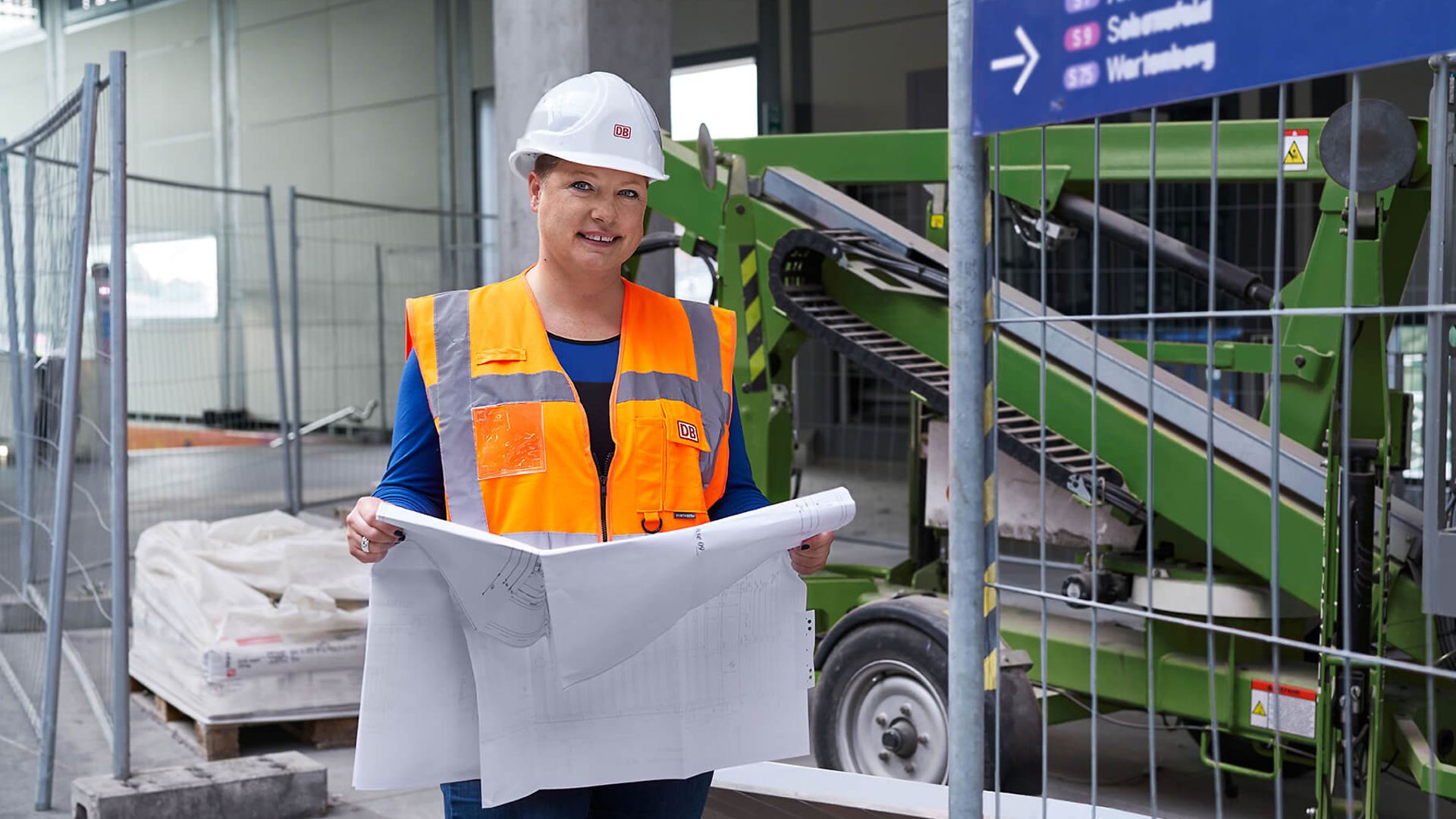 Eine Frau mit Schutzweste und Helm hält ein großen Plan vor sich. Sie steht vor einer Baustelle an einem Bahnhof.
