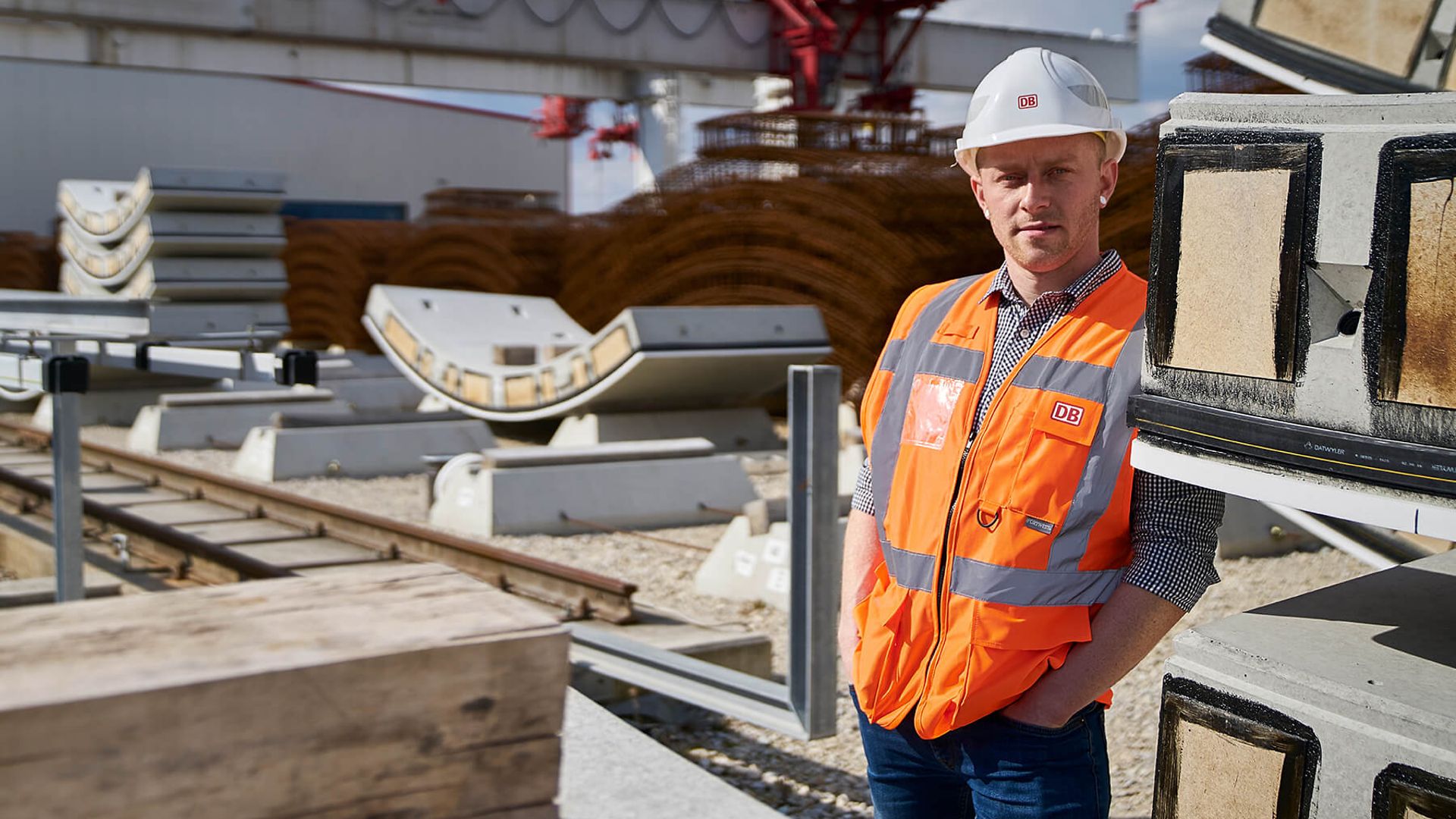 Ein Mann mit Warnweste und Helm blickt in die Kamera. Neben ihm liegen Gleise und viele andere Materialien, wie Holz und Metall.