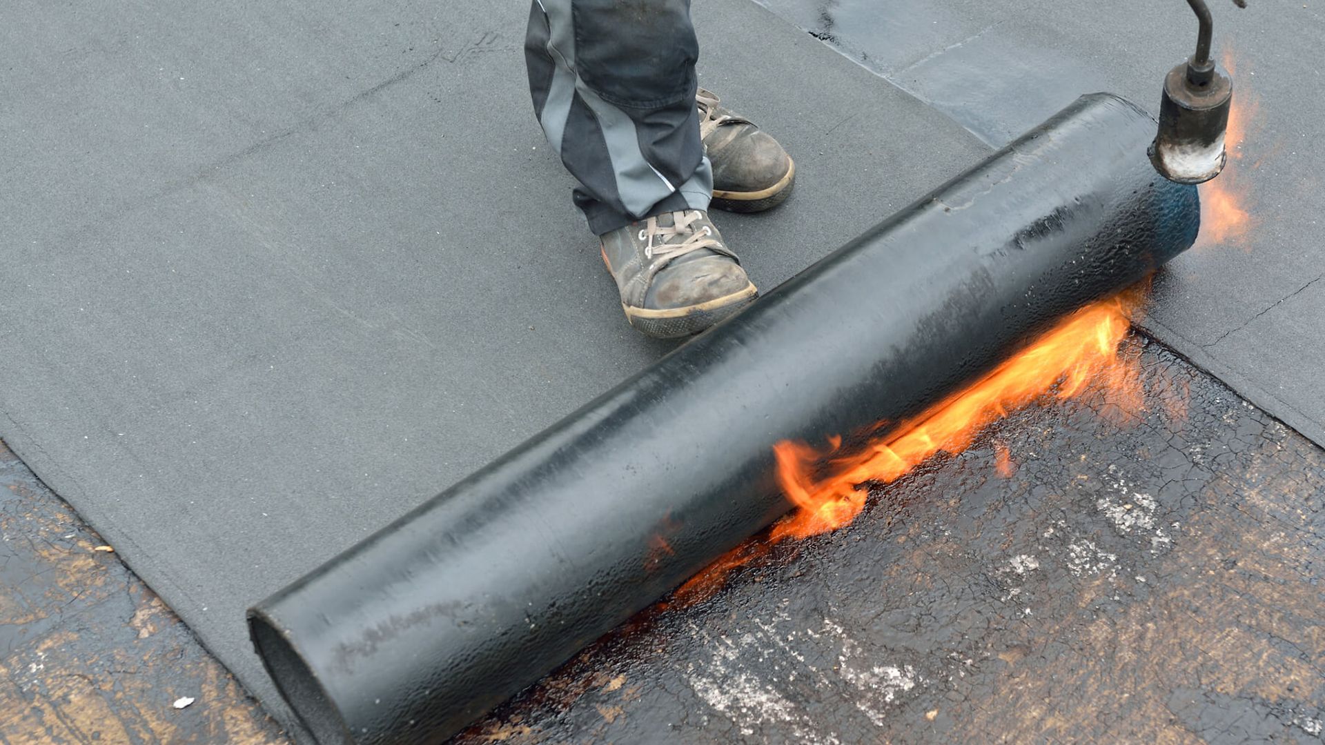 Detailaufnahme einer Dachpappe, die mit einem Gasbrenner erhitzt und ausgerollt wird.