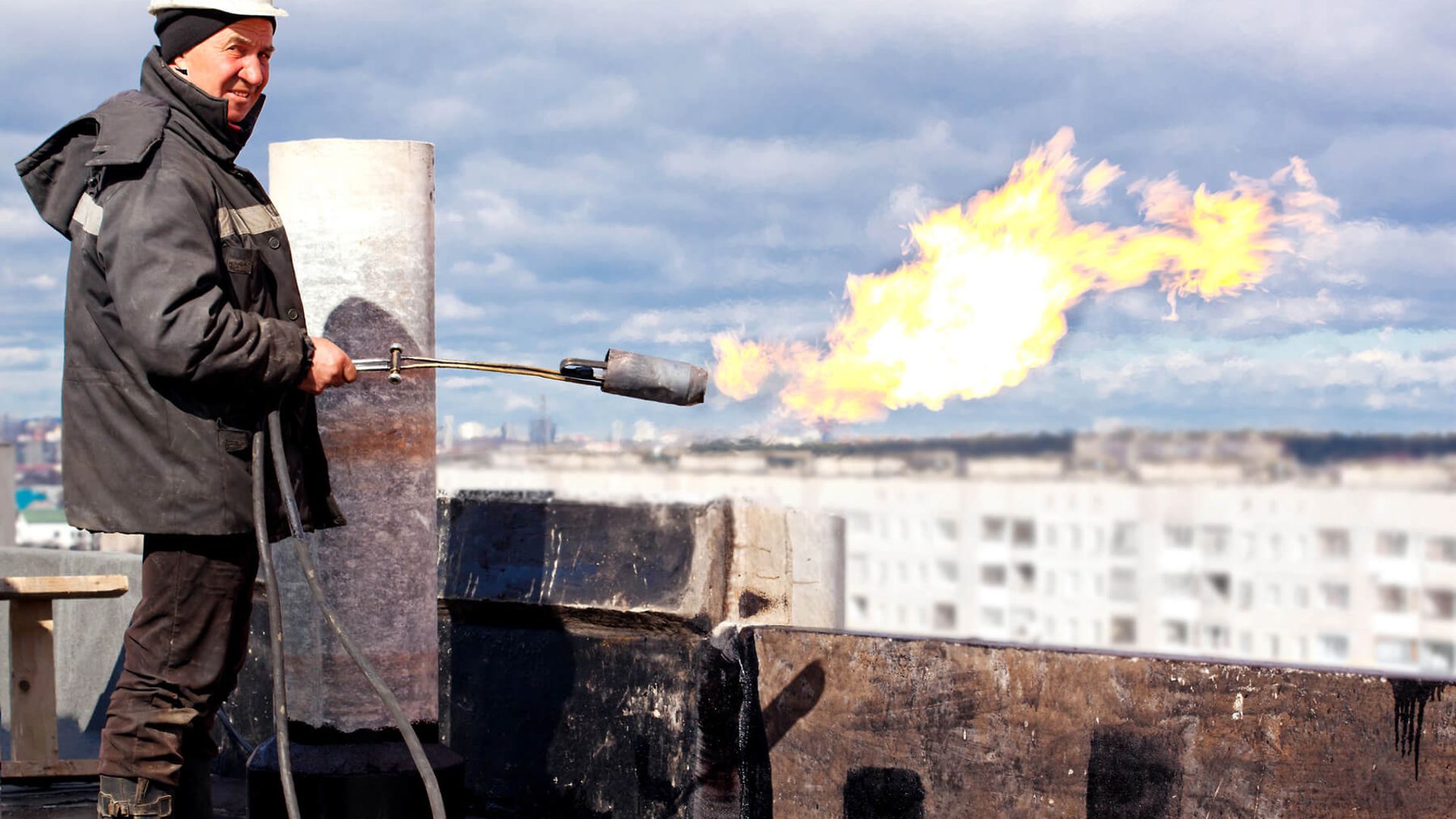 Eine Person mit Helm und Schutzkleidung hält einen Gasbrenner der gerade eine Flamme erzeugt und steht auf einem Hausdach.