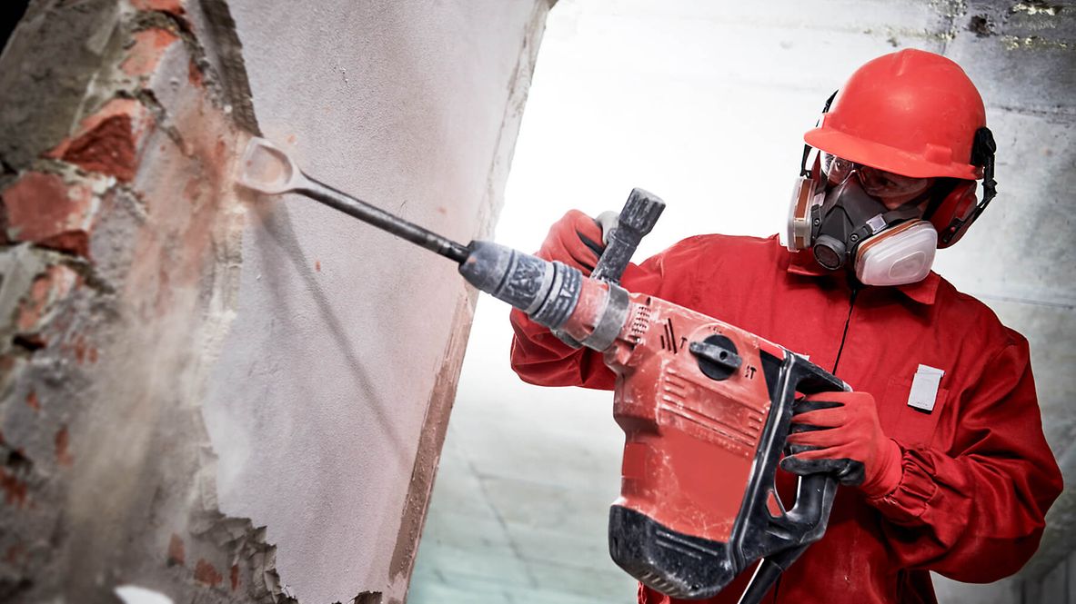 Eine Person steht mit voller Schutzkleidung an einer Wand und bohrt.