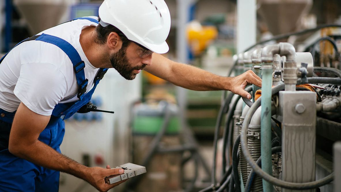 Ein Mann in Latzhose und Schutzhelm betrachtet intensiv eine Maschine.