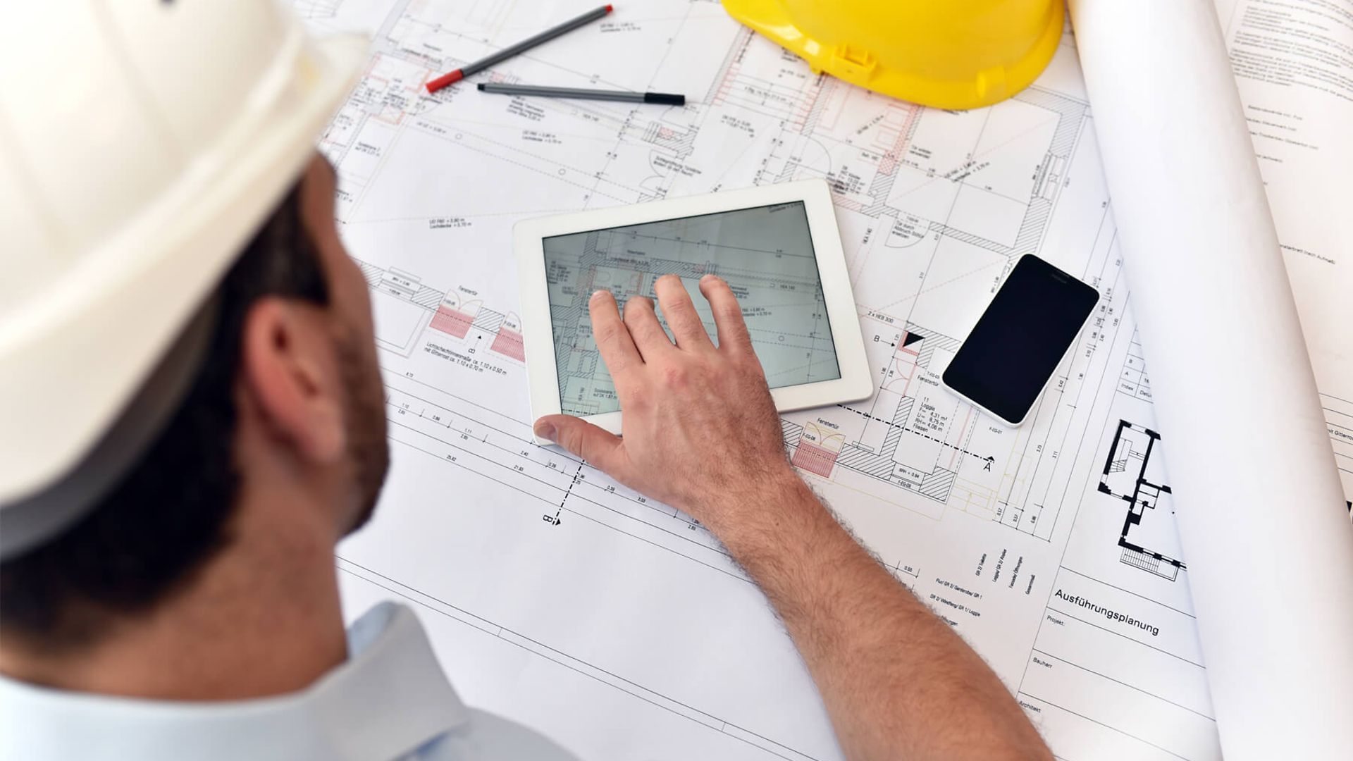 Ein Mann tippt auf seinem Tablet. Das Tablet liegt auf Plänen von technischen Zeichnungen.