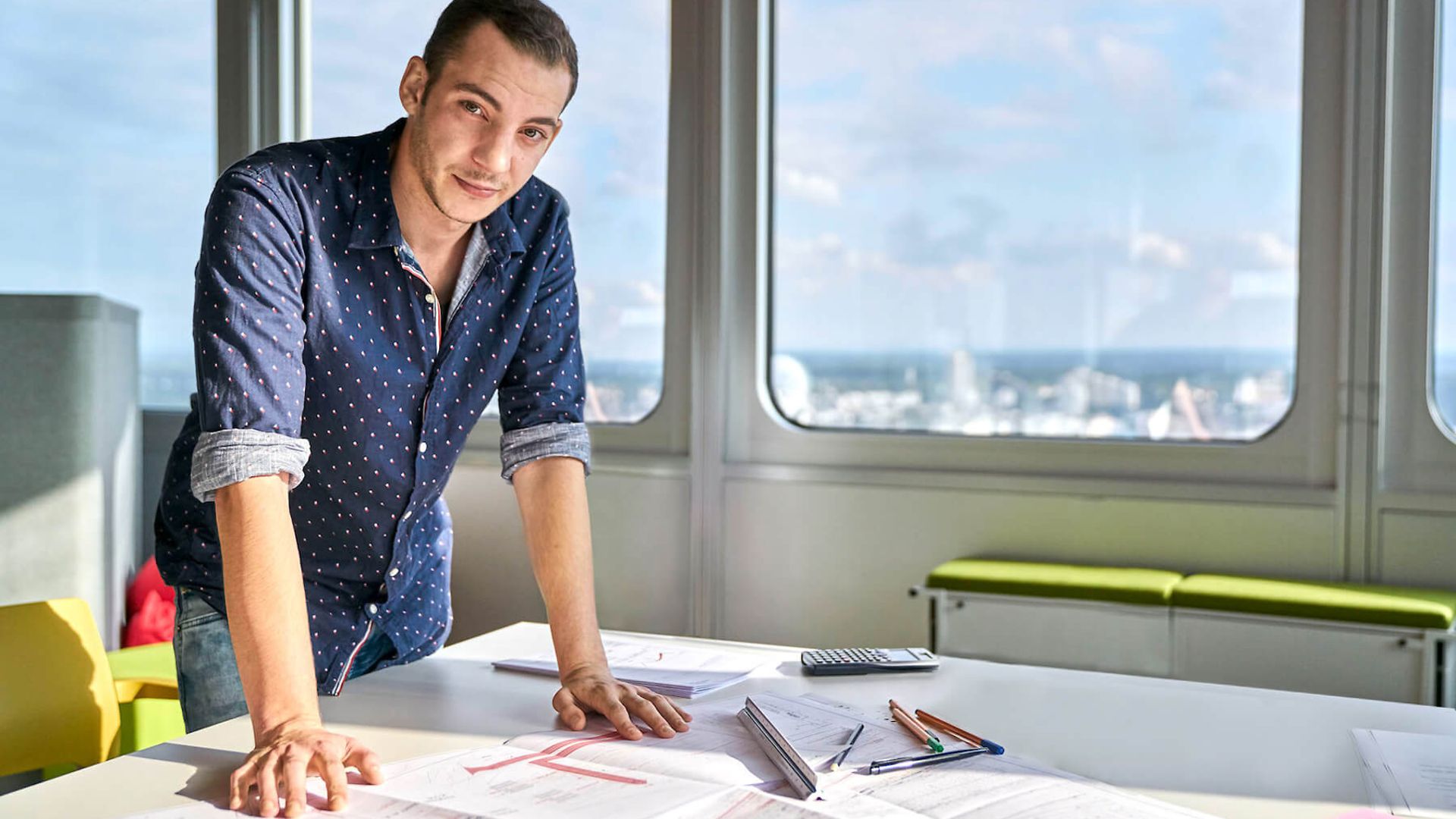 Ein Mann steht an einem großen Tisch in einem Büroraum und stützt die Arme auf, dabei sieht er in die Kamera. 