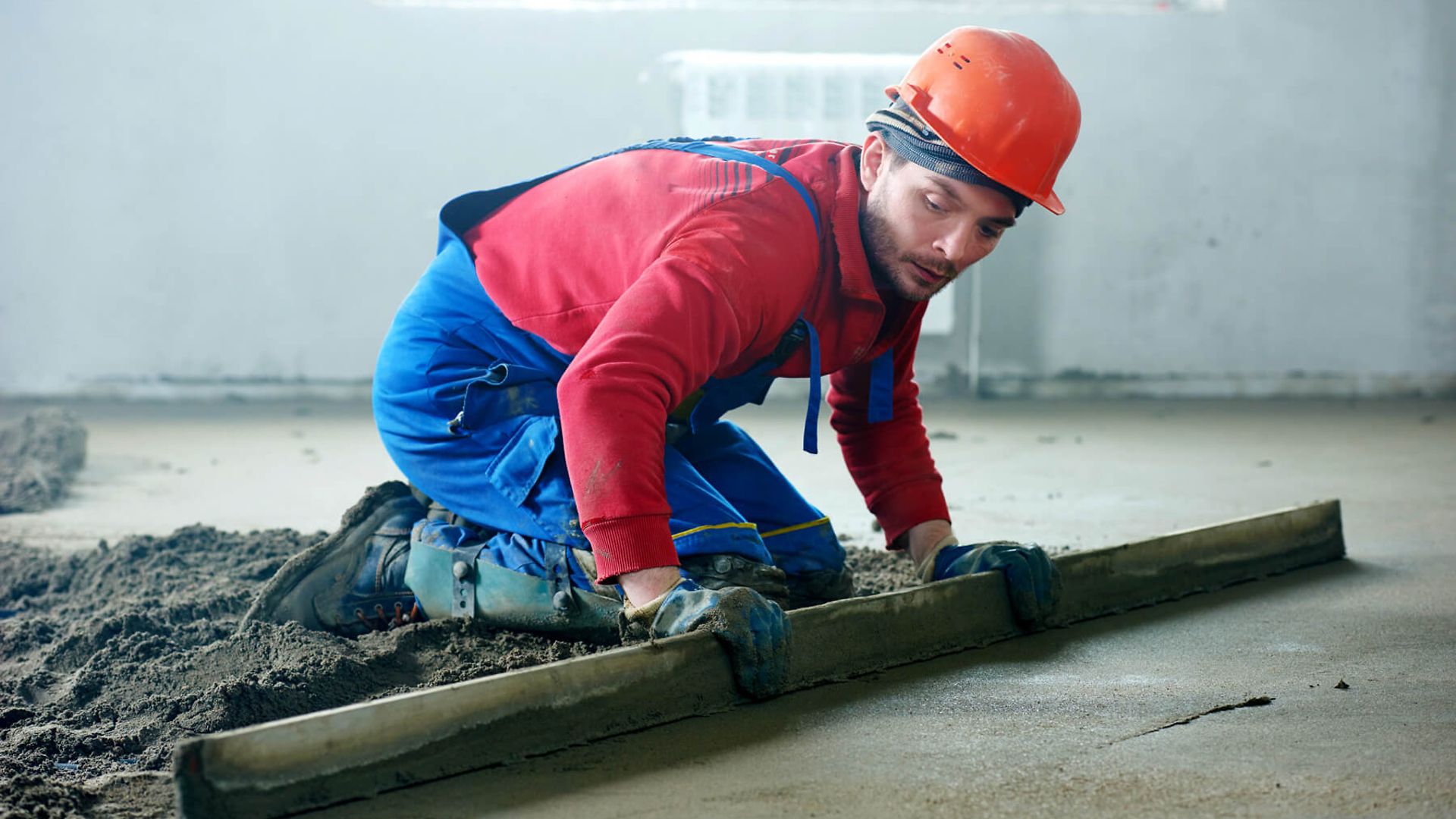 Ein Mann streicht Beton glatt. Er hat eine blaue Latzhose und einen Roten Helm an. Für die Arbeit kniet er auf dem Boden.