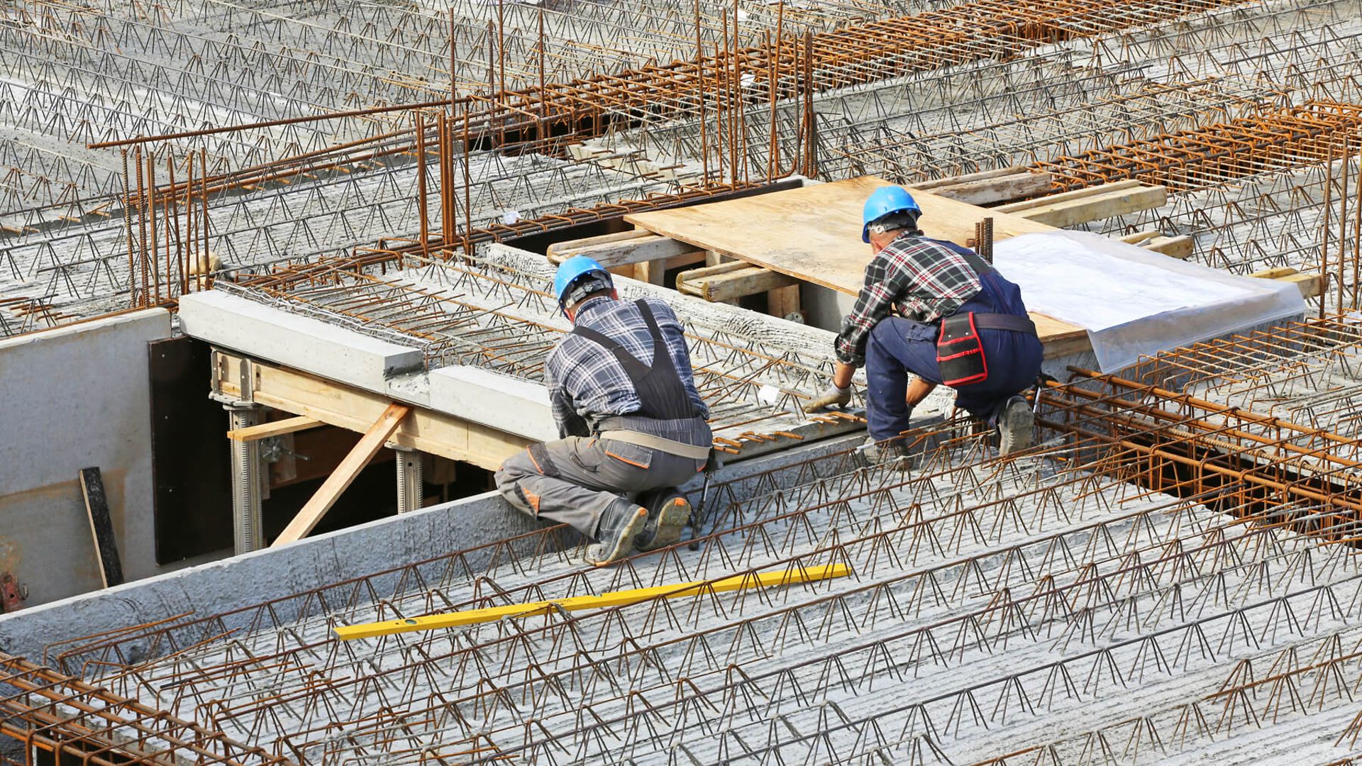 Zwei Personen in Arbeitskleidung mit Schutzhelm knien auf einem Rohbau. Es sind Halbfertigteildecken von oben zu sehen. 