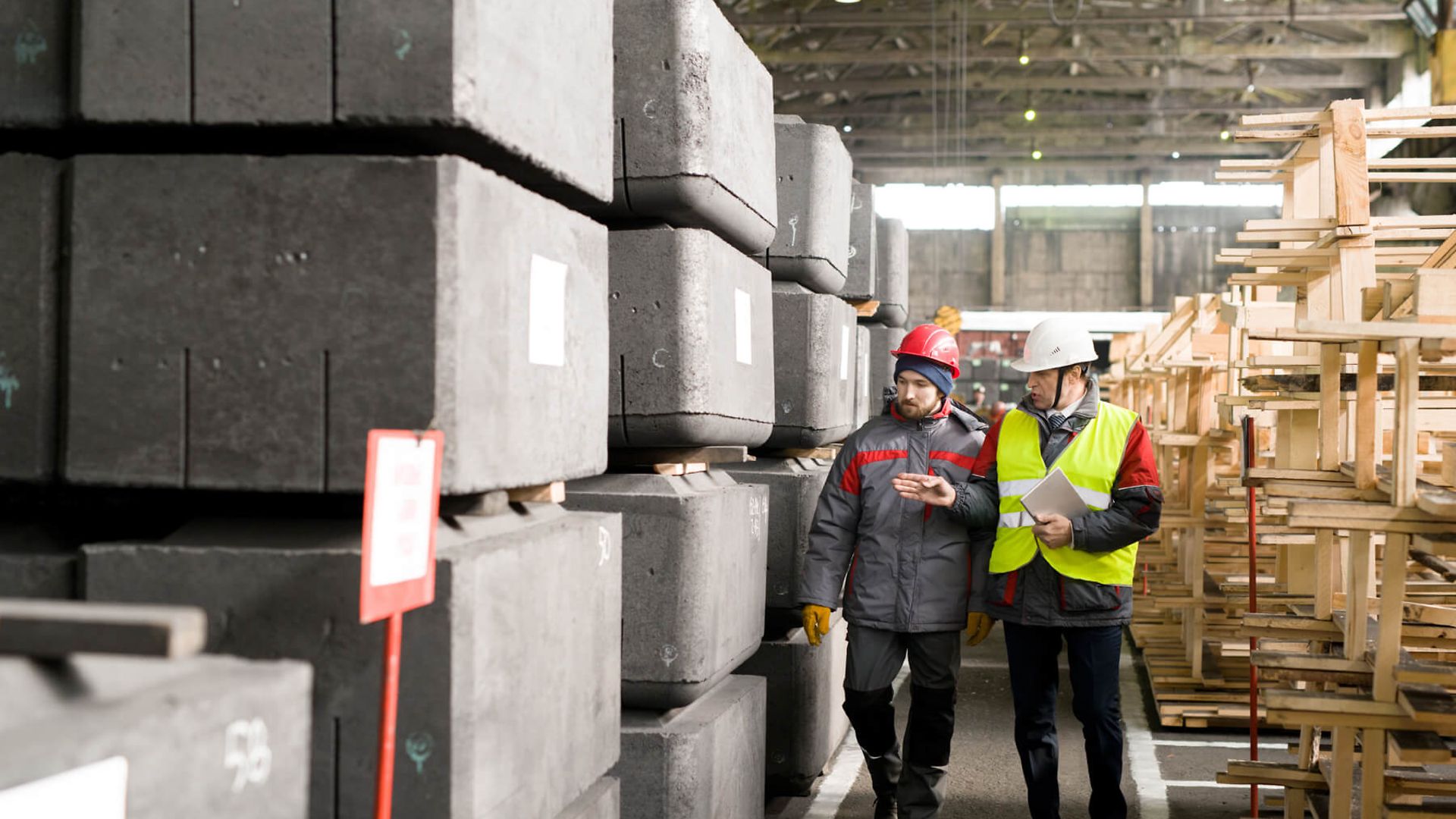 Zwei Männer in Arbeitskleidung mit Schutzhelm laufen durch ein Lager mit Baumaterial zwischen Holzstapeln und Betonklötzen. 
