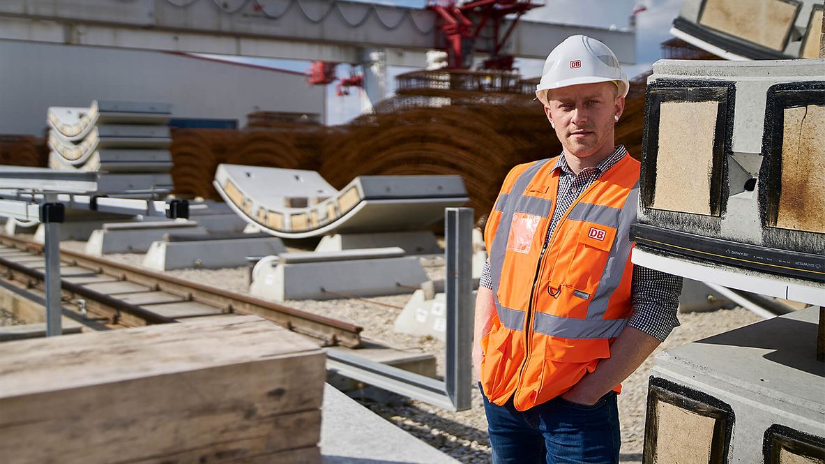 Ein Mann mit Warnweste und Helm blickt in die Kamera. Neben ihm liegen Gleise und viele andere Materialien, wie Holz und Metall.