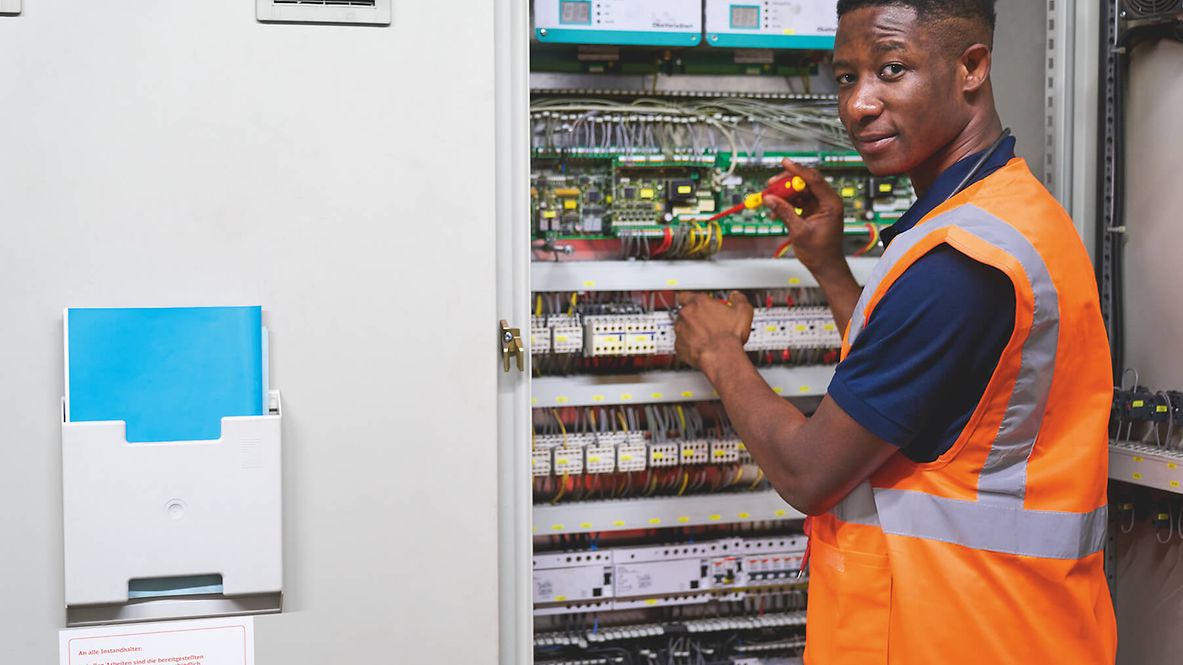 Ein Mann steht vor einem offenen Sicherungskasten und hält einen Phasenprüfer, dabei dreht er sich um und blickt in die Kamera.