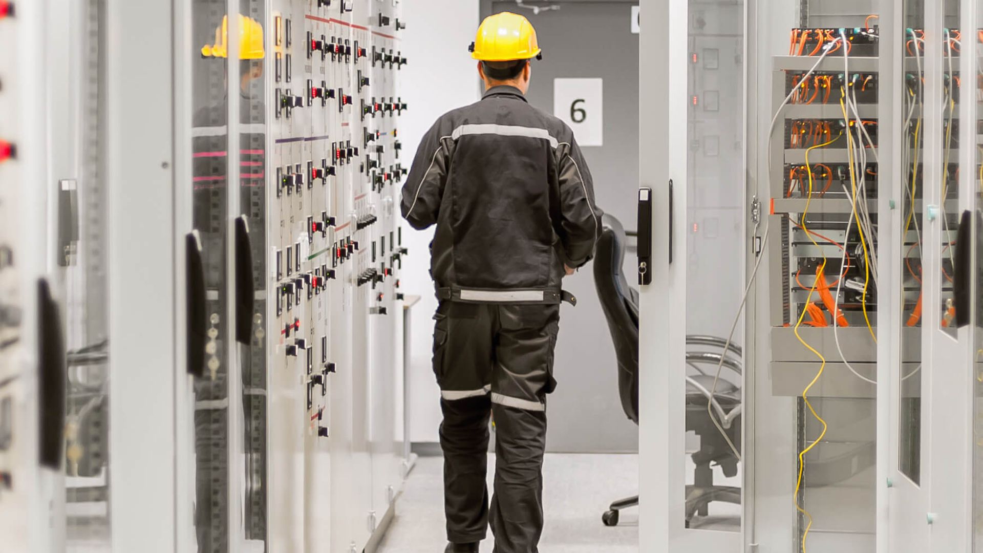 Eine Person geht von der Kamera weg. Er geht durch den Gang eines Elektroraumes. Es sind viele Schränke mit Kabeln zu sehen.