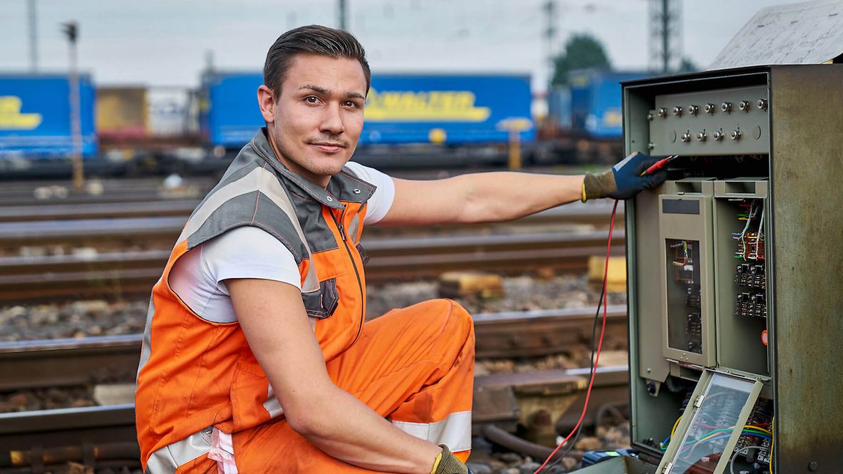 Ein Mann kniet vor einem Kasten im Außenbereich der Gleise und dreht sich zur Kamera.
