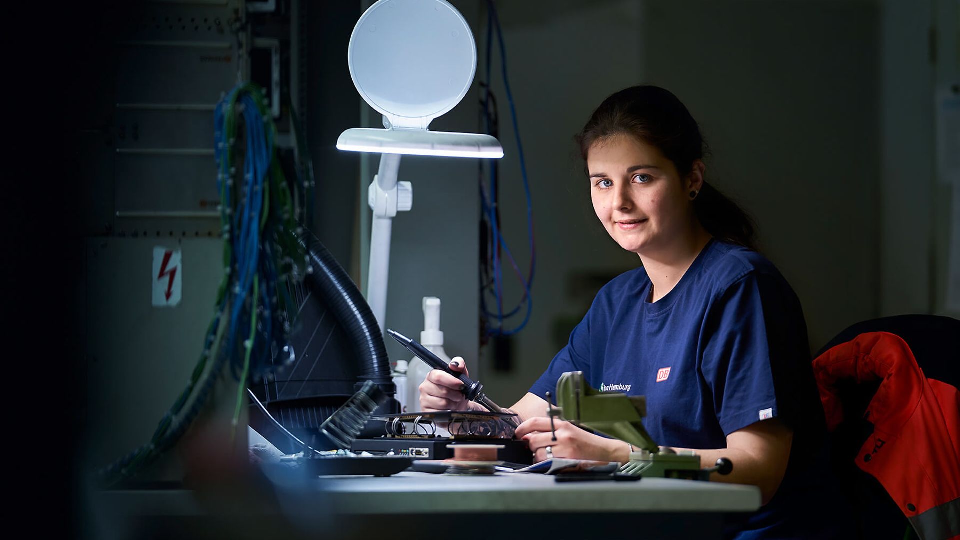Eine Frau sitzt an einem Schreibtisch mit mehreren kleinen Werkzeugen und einer hellen Arbeitslampe
