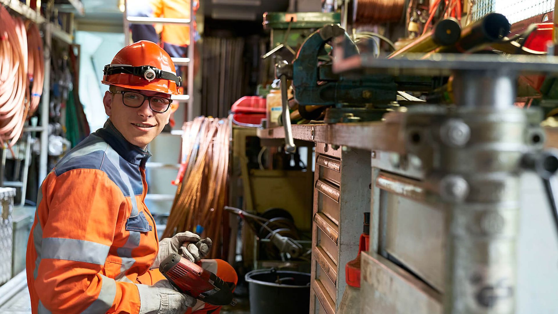 Ein Mann kniet vor einer Werkbank in einer großen Werkstatt. Erhält einen Akkuschrauber in der Hand.