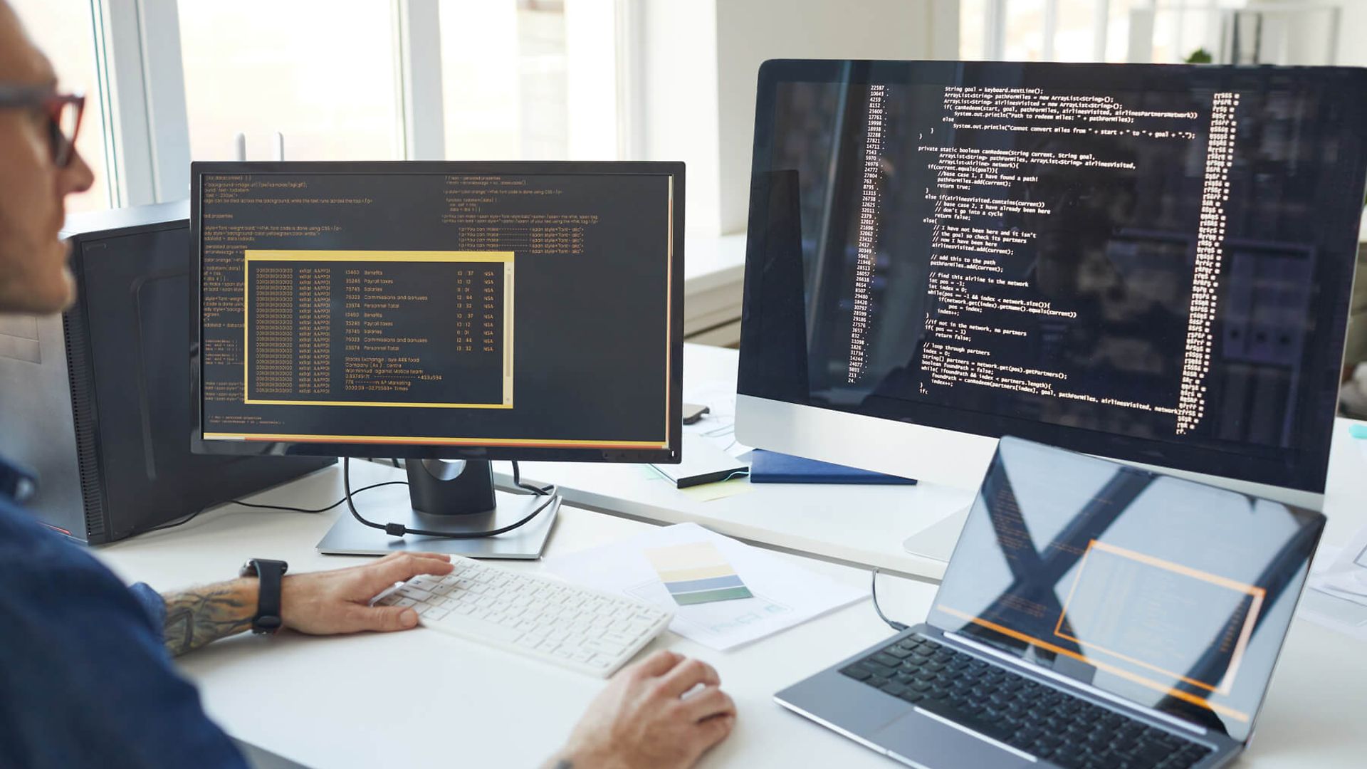 Ein Mann sitzt an einem Schreibtisch. Vor ihm stehen 2 Bildschirme. Auf den Bildschirmen sind Codes zu sehen.