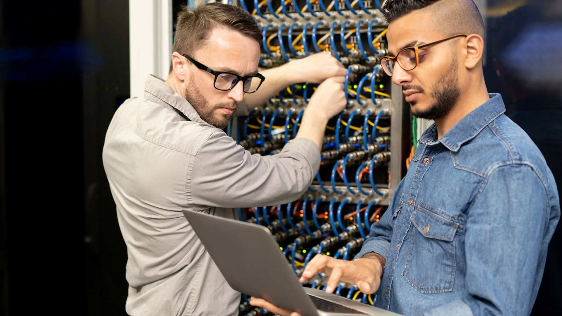 Zwei Männer, der Mann im Vordergrund hat einen Laptop in der Hand der andere steht an einem Kasten mit vielen Kabeln