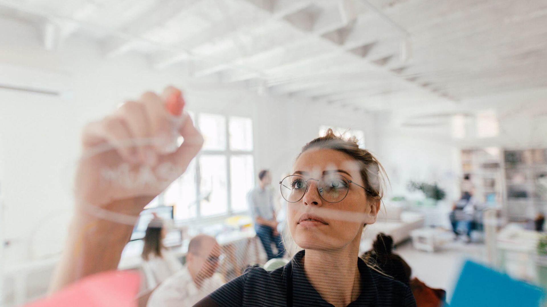Eine Frau schreibt mit einem weißen Marker auf eine Glaswand. Der Blick ist durch die Glaswand auf die Frau gerichtet