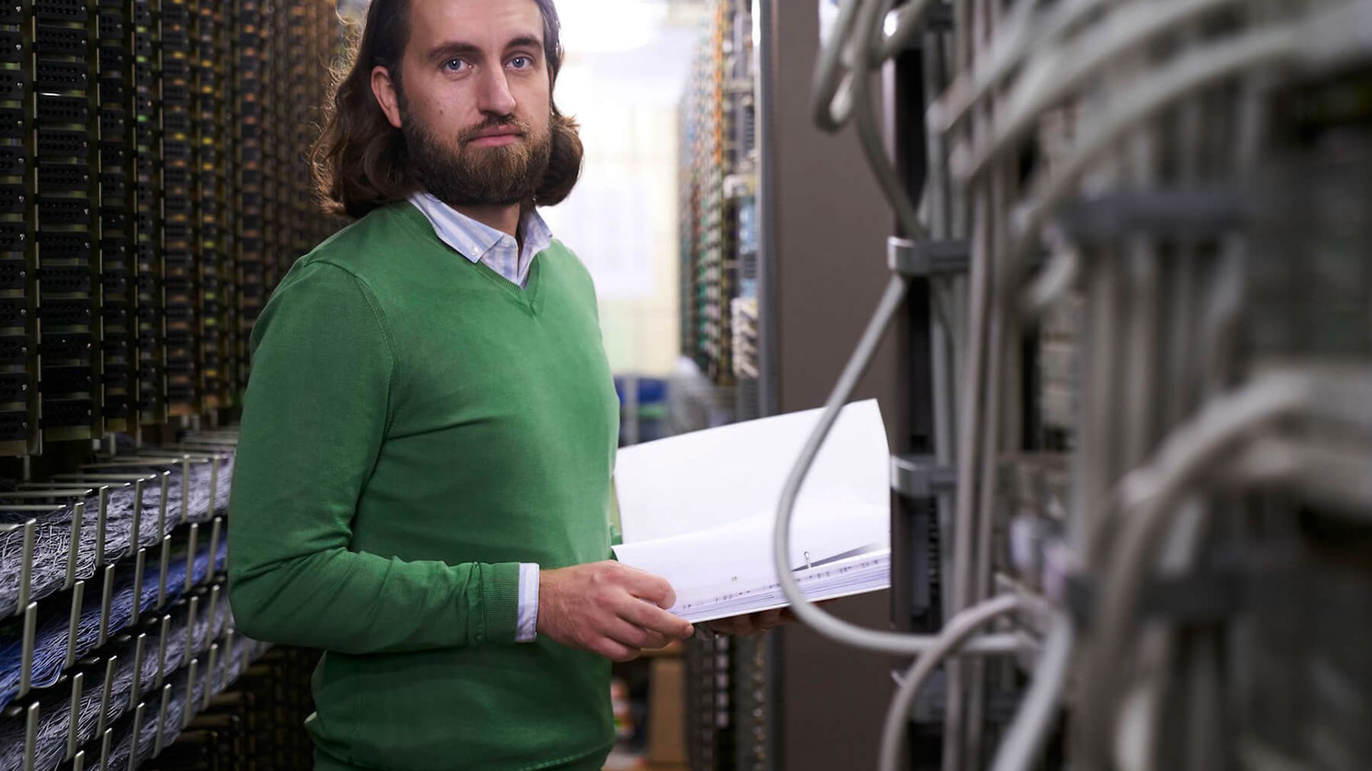 Ein Mann steht in einem Gang zwischen Servern. Er schaut direkt in die Kamera und hat ein Buch in der Hand.