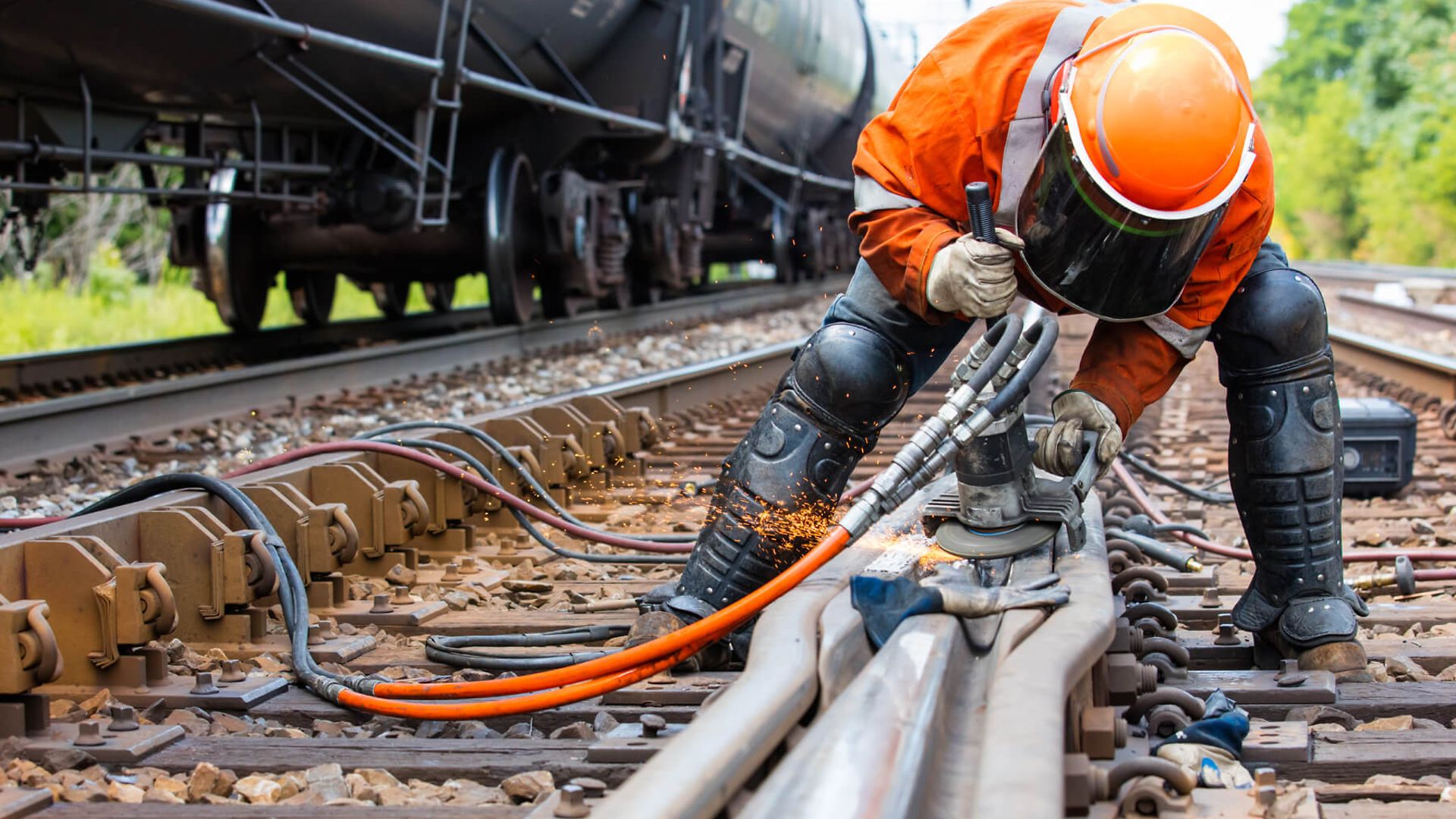 Eine Person arbeitet mit einer Maschine am Gleis. Dazu steht er mit Schutzkleidung und Visier über das Gleis gebeugt.