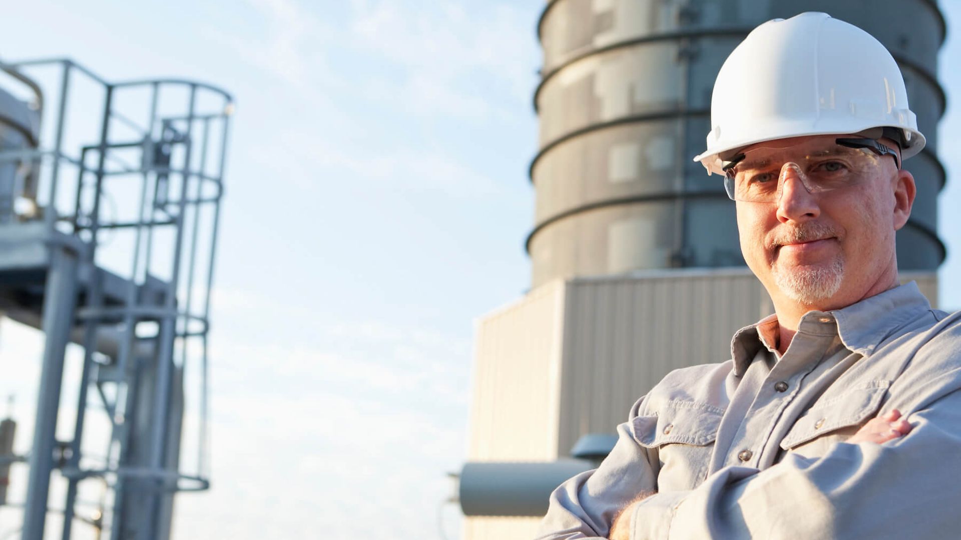 Ein Mann mit Helm und Schutzbrille blickt in die Kamera. Im Hintergrund ist ein fabrikartiges Gebäude zu sehen.