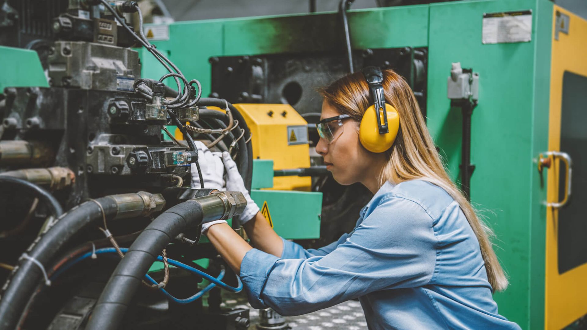 Eine Frau mit Handschuhen und Gehörschutz arbeitet an einer großen Maschine mit vielen Kabeln.