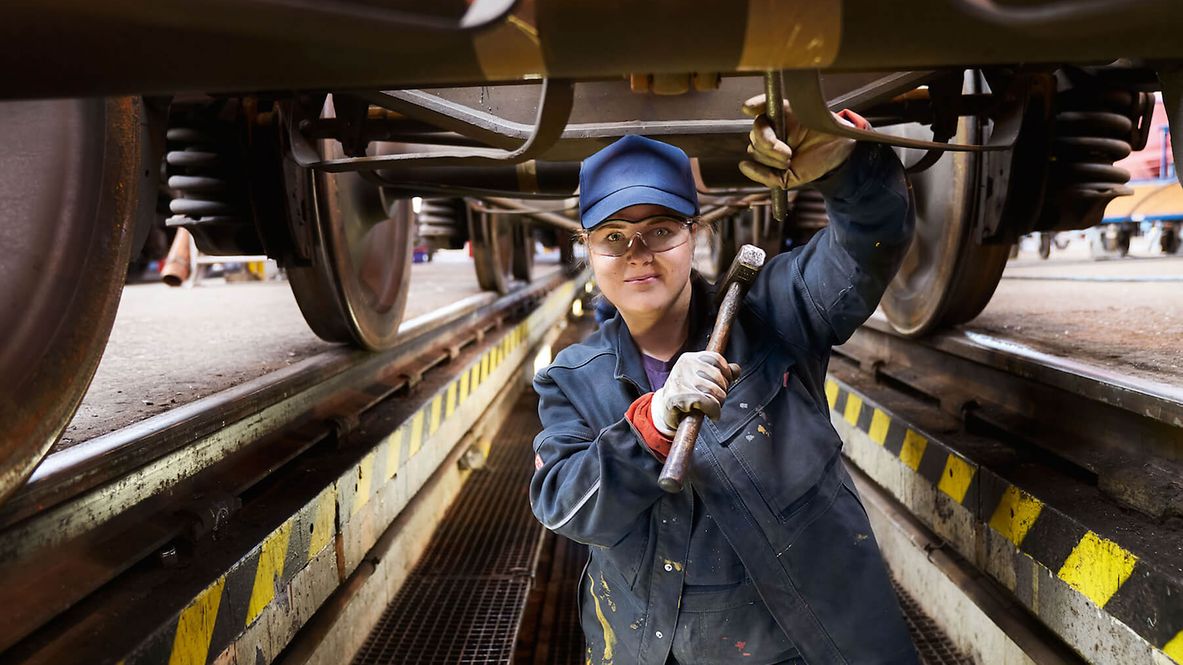 Eine Frau steht mit stark verschmutzter Arbeitskleidung unter einem Zug. In der Hand hält sie einen Metallhammer.