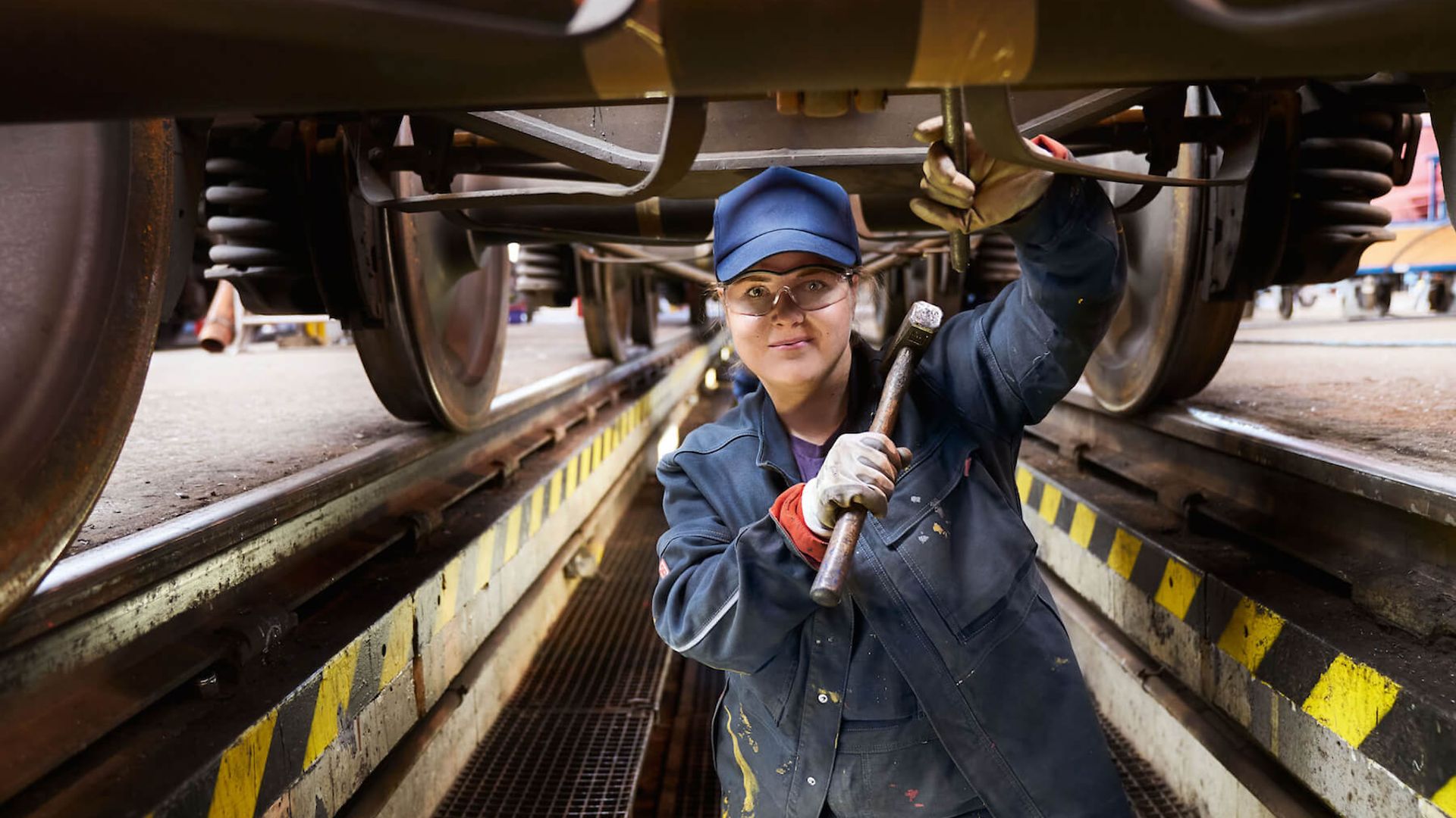 Eine Frau steht mit stark verschmutzter Arbeitskleidung unter einem Zug. In der Hand hält sie einen Metallhammer.