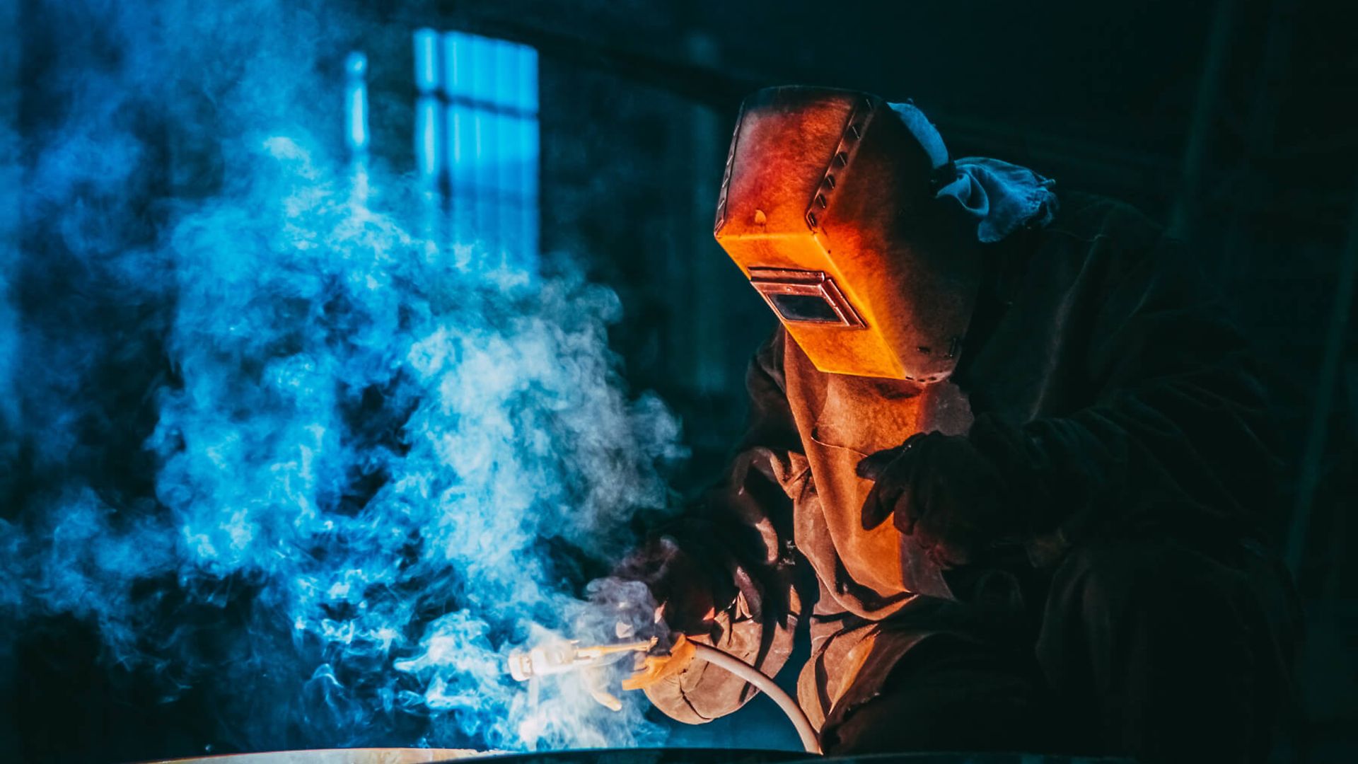 Eine Person mit voller Schutzkleidung arbeitet mit einem Schweißgerät. Es ist eine blaue Flamme und Rauch zu sehen.