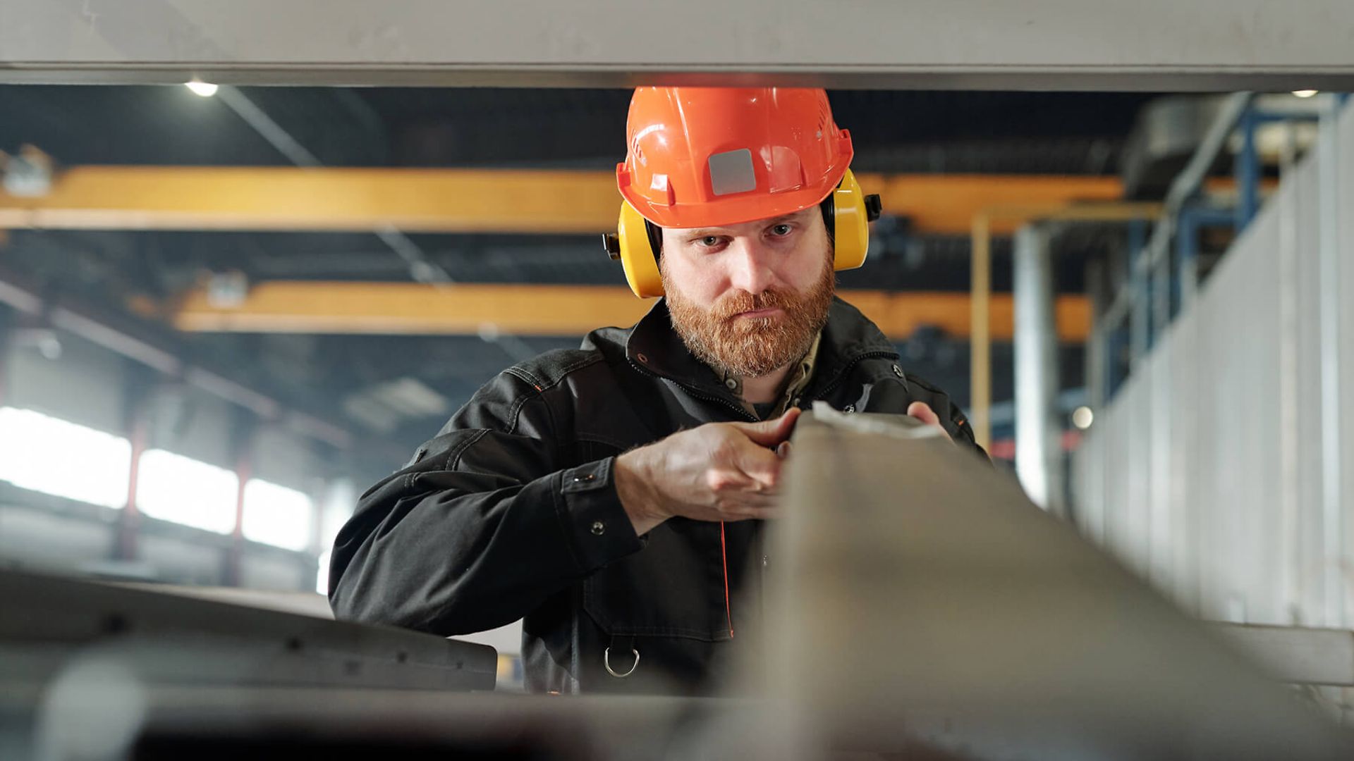 Ein Mann mit orangenem Helm und gelbem Gehörschutz hält eine Metalllatte.