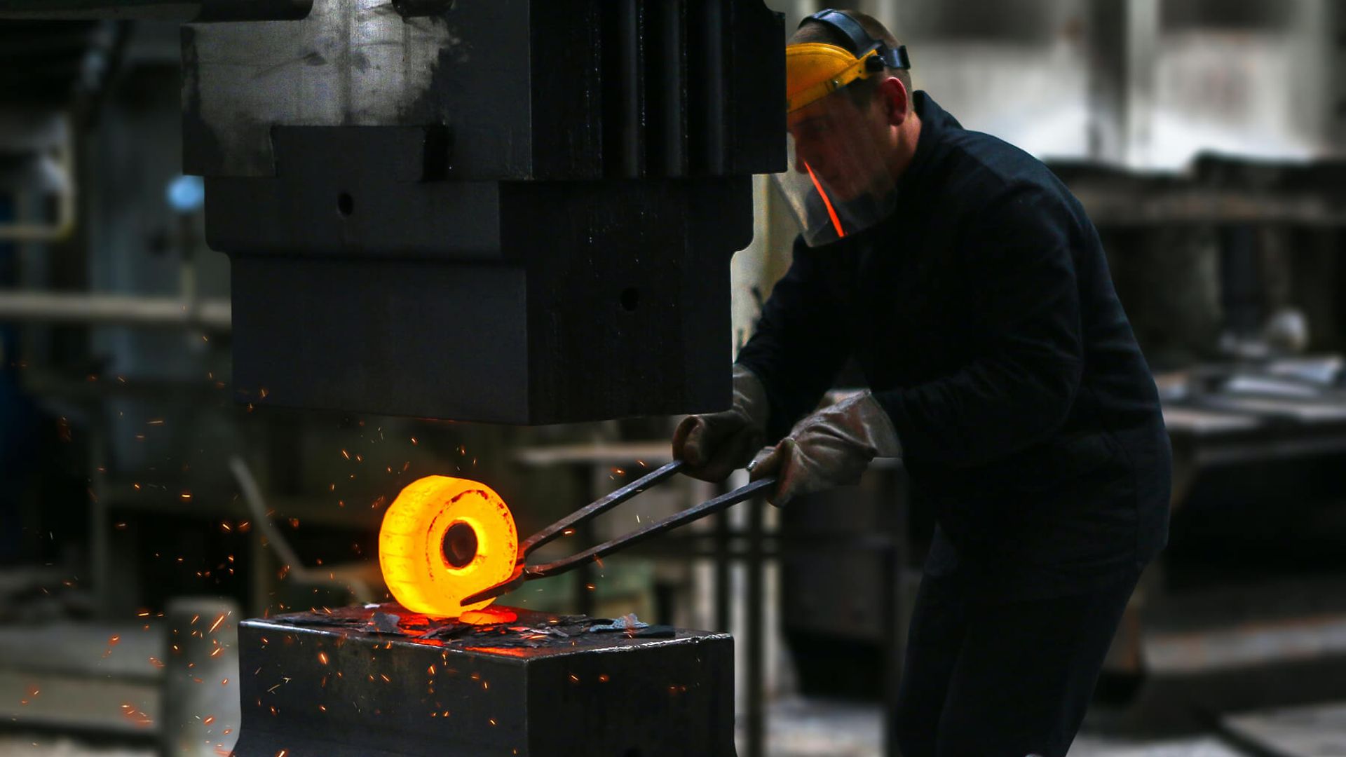 Eine Person hält mit einer langen Zange ein glühendes Metallteil unter einen Amboss. Die Person trägt einen Schutzanzug.