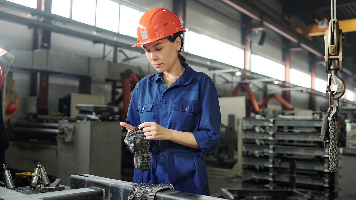 Eine Frau steht mit Arbeitskleidung und Helm vor einem Metaltisch in einer Werkstatt und zieht sich ihre Arbeitshandschuhe an.