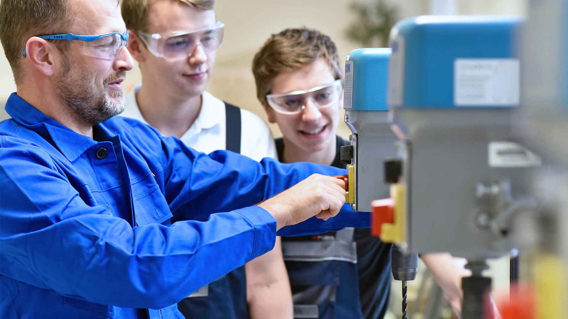 Ein Mann mit Blaumann und Schutzbrille bedient eine Maschine. Zwei jüngere Männer sehen ihm dabei zu.