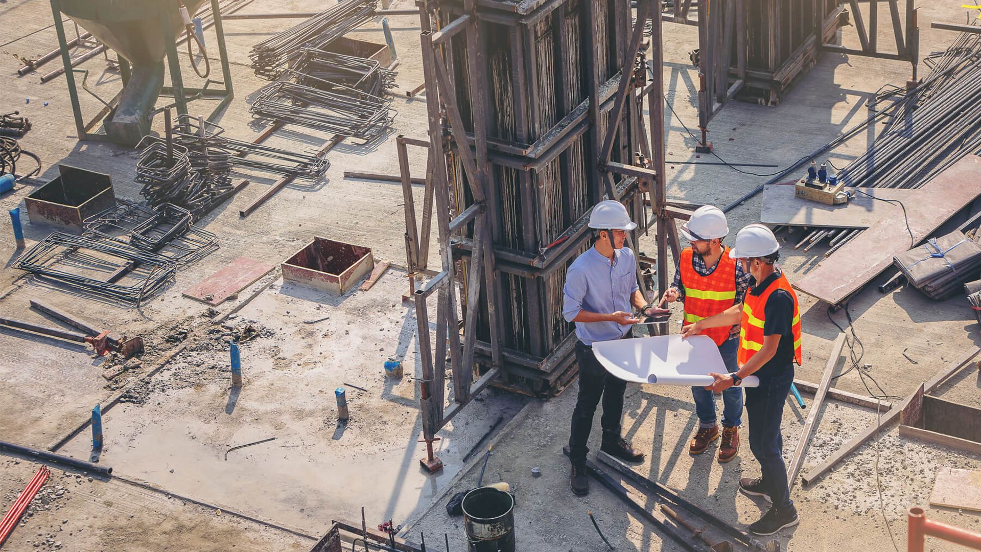 3 Personen stehen mit Warnweste und Helmen auf einem Rohbau, sie halten ein Plan in der Hand.