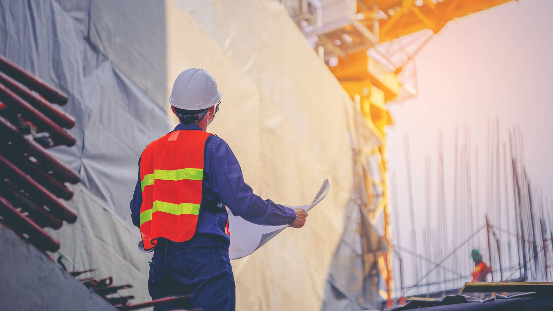 Eine Person mit Warnweste und Helm steht mit einem Plan in der Hand vor einer großen Baustelle.
