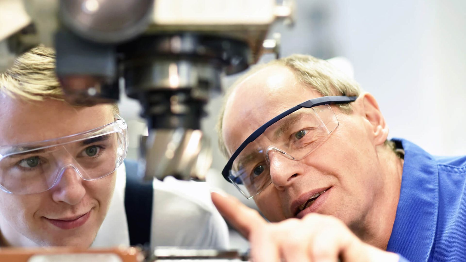 Eine Person erklärt einer anderen Person eine Maschine. Beide tragen Blaumann und Schutzbrille.