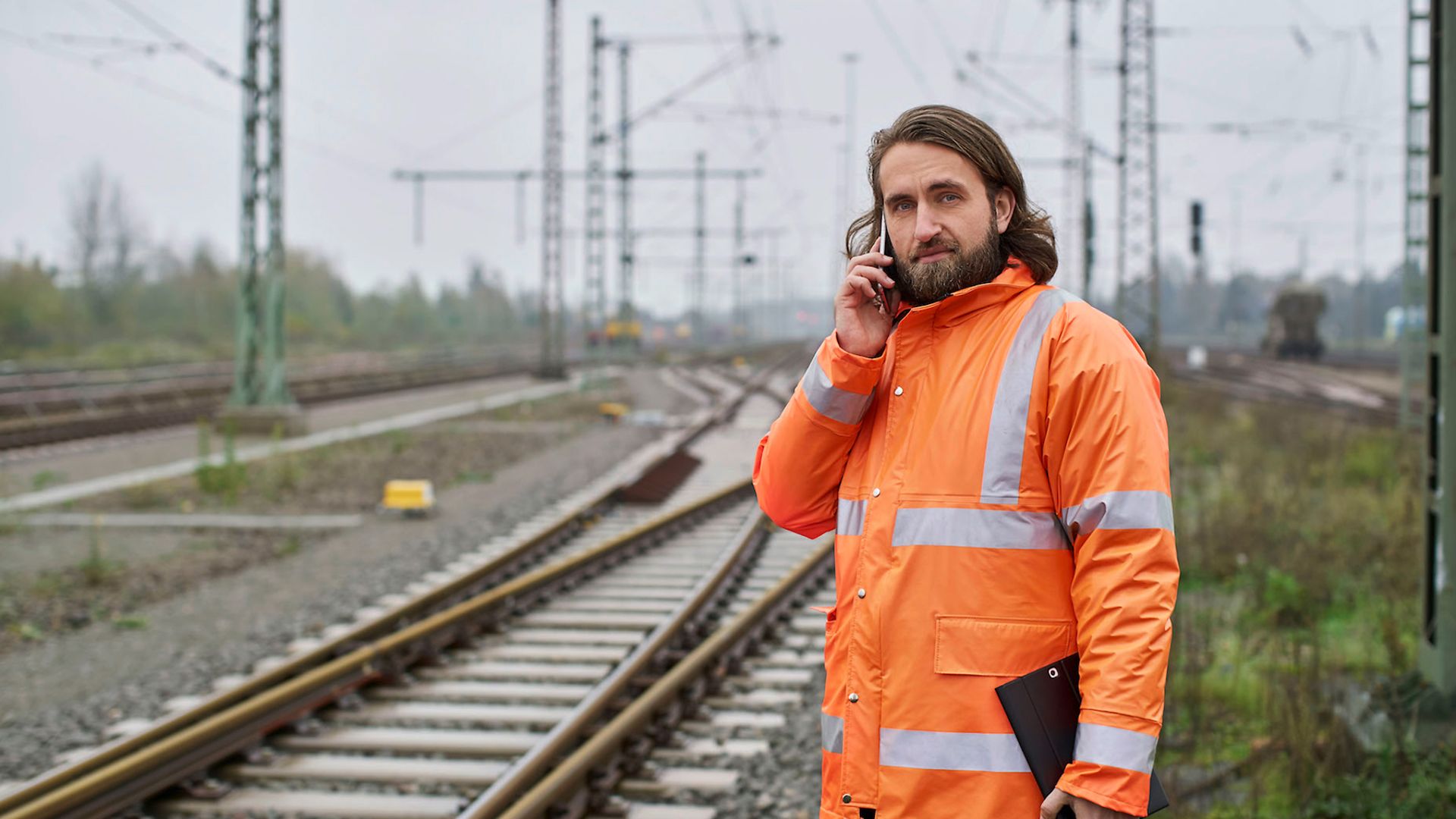 Ein Mann in einer orangenen Warnjacke steht an der Gleisstrecke. Er telefoniert mit dem Handy