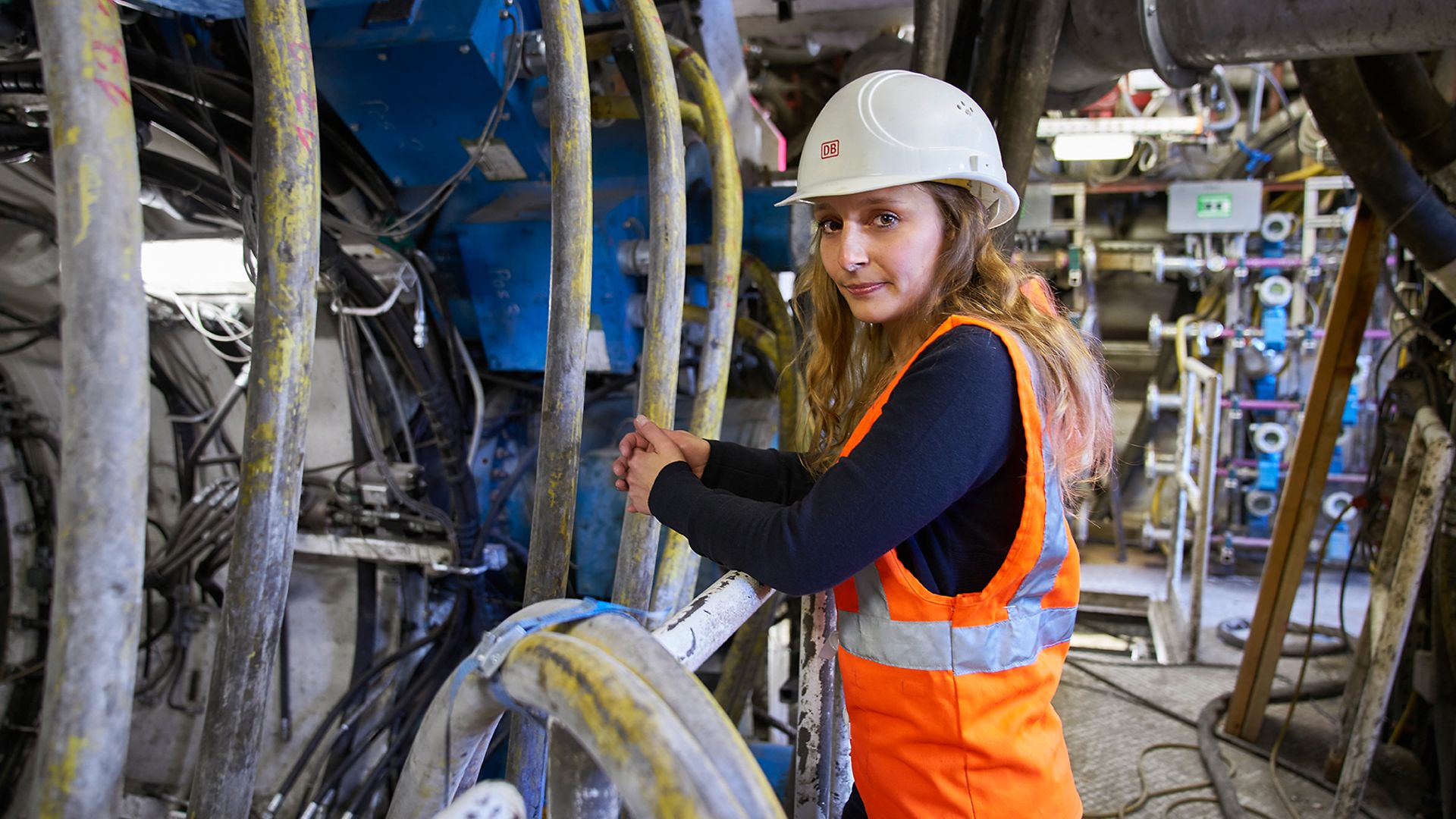Eine Frau mit Warnweste und Helm steht in einem Raum mit Leitungen, Maschinen und Rohren.