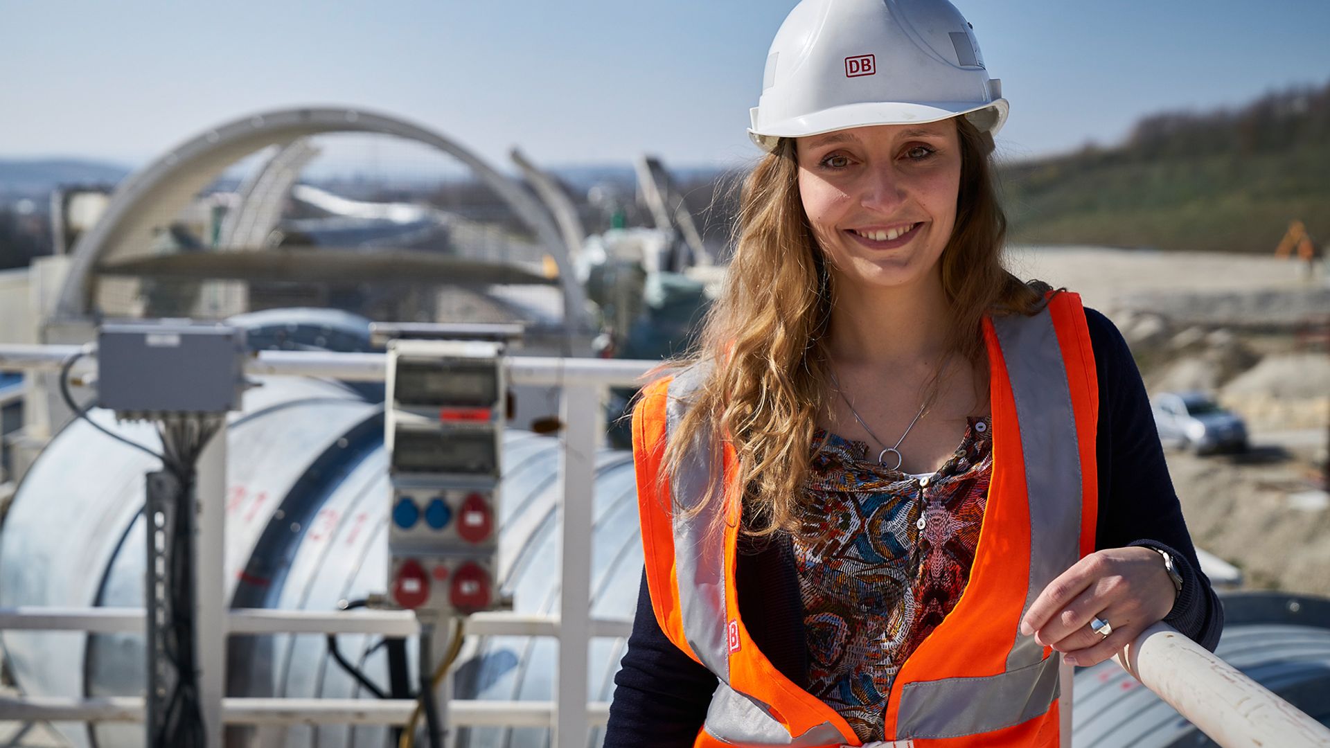 Eine Frau mit Warnweste und Helm lächelt in die Kamera. Im Hintergrund sind sehr große Rohe in einer Baugrube zu erkennen.